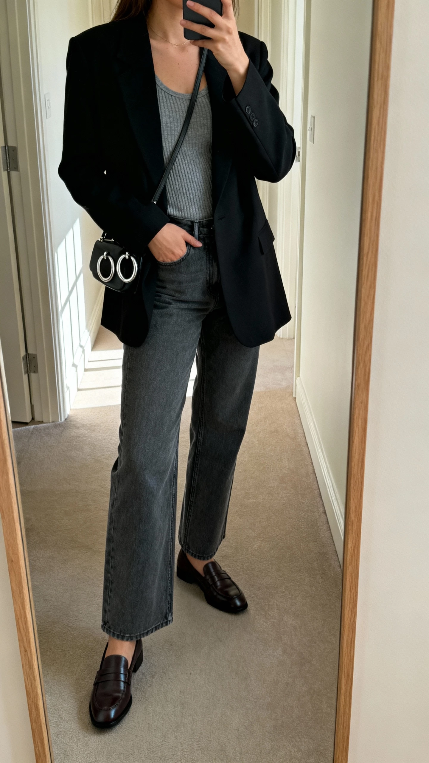 iPhone side-angle mirror selfie of a woman wearing charcoal bootcut jeans, a minimalist black tailored blazer over a gray ribbed tank, sleek leather loafers, and a small crossbody bag with silver hoops; face not visible, hallway mirror with morning window light, natural iPhone photo quality.