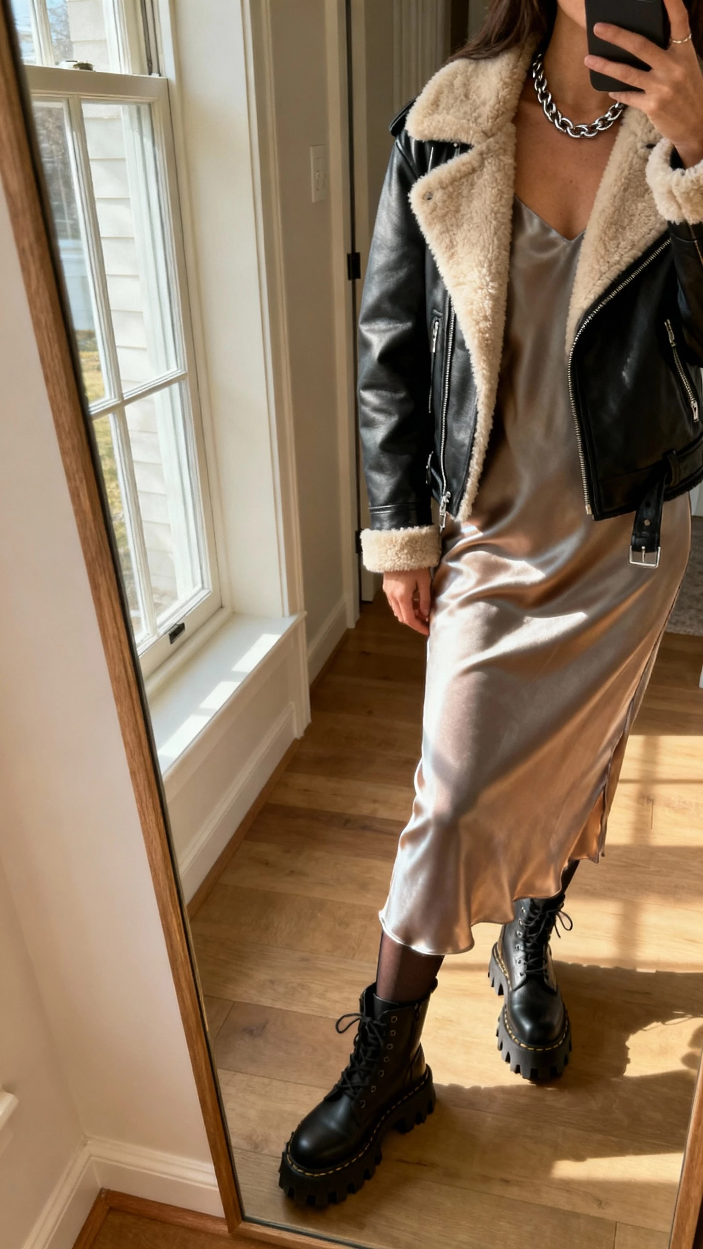 iPhone side-angle mirror selfie of a woman wearing a shearling-lined moto jacket over a satin slip dress, opaque tights, lug-sole boots, chain necklace, face not visible, entryway mirror, late-afternoon window light, iPhone photo