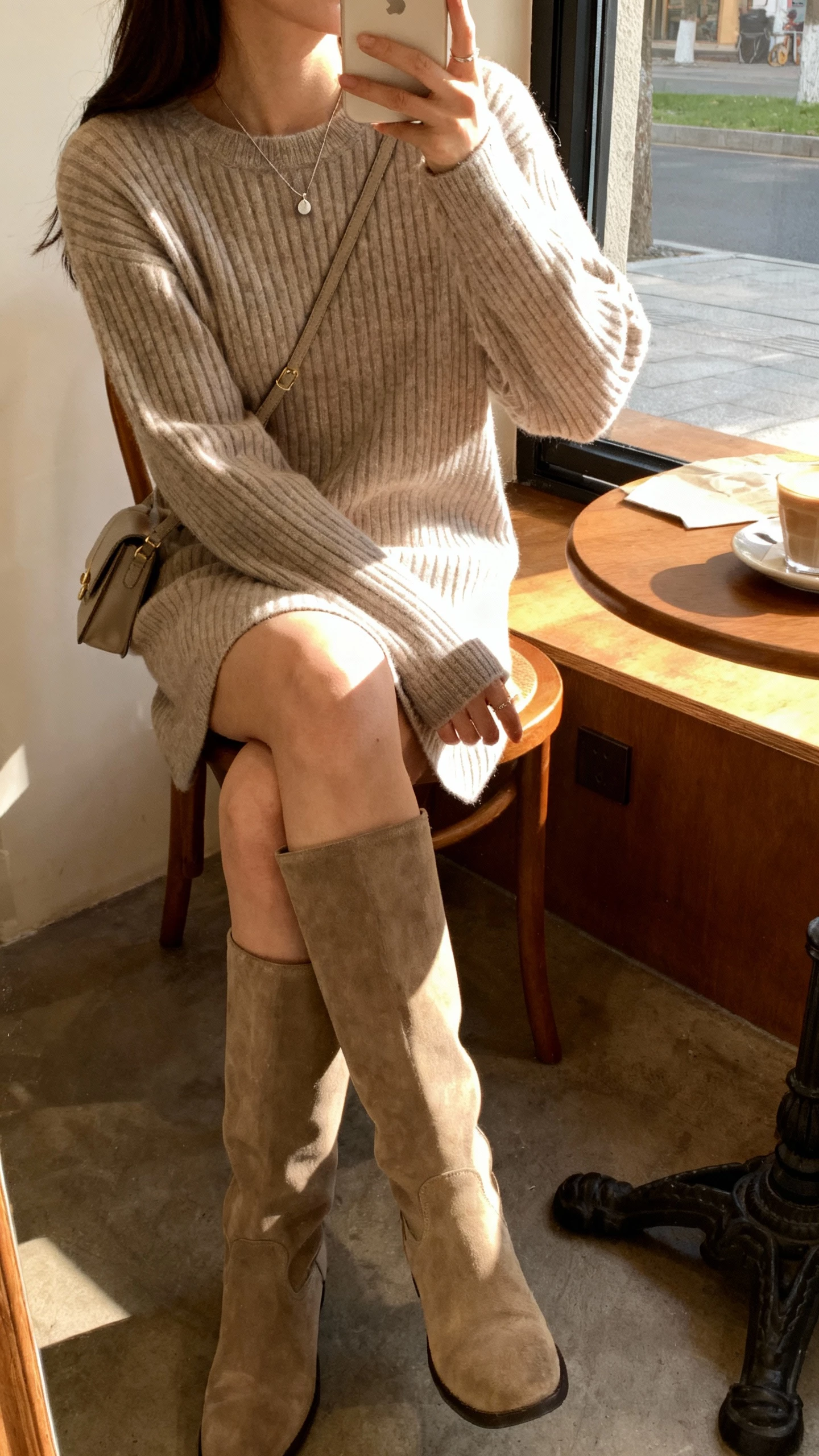 iPhone side-angle seated selfie of a woman in a ribbed sweater dress with tall boots, minimal pendant necklace, and a small shoulder bag, face not visible, cozy cafe corner by the window, warm morning light, natural iPhone photo quality