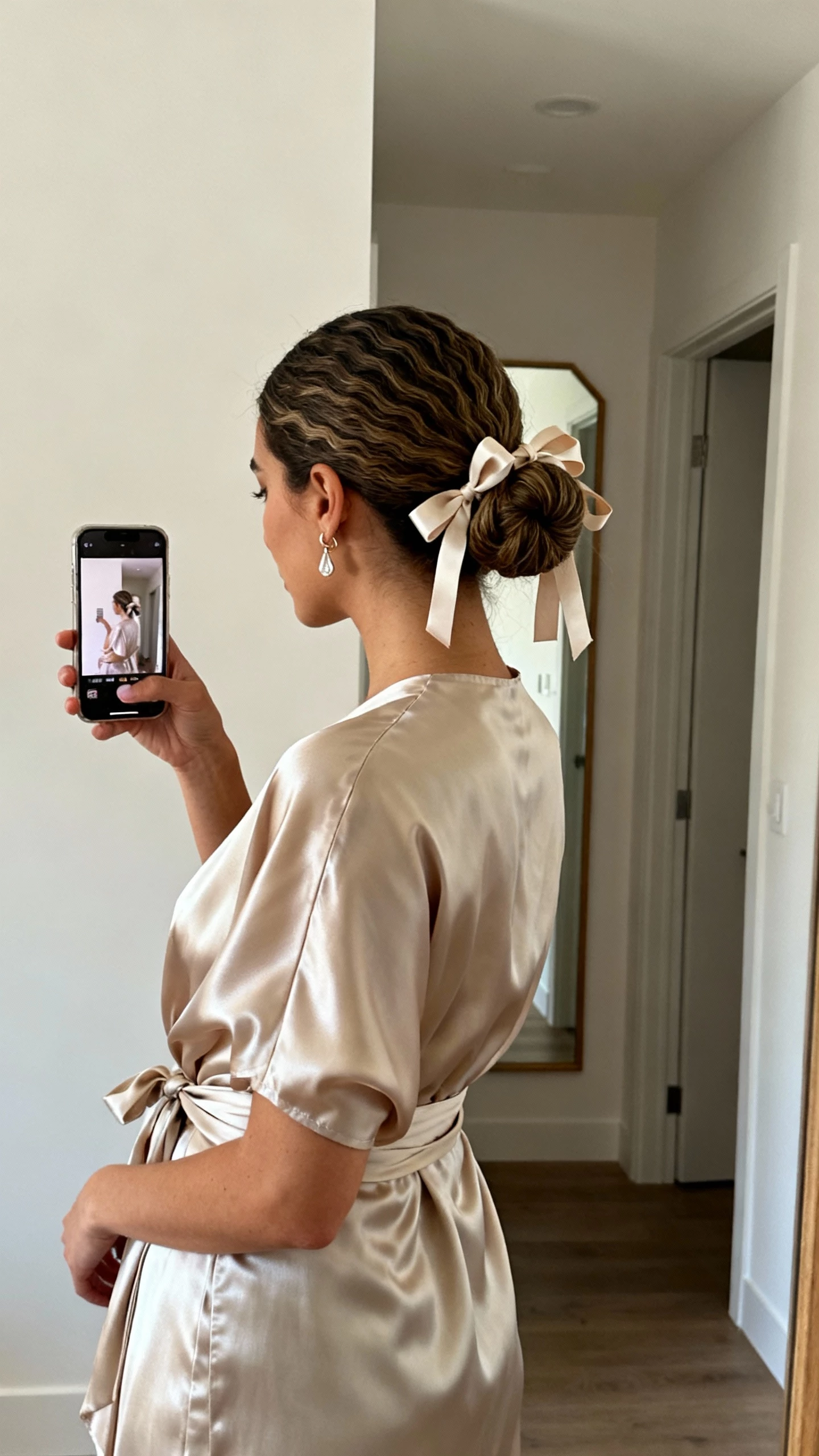 iPhone side-angle selfie capturing a low textured chignon tied with soft ribbon bows, a flowy satin wrap dress, dainty drop earrings, face not visible, hallway mirror in soft indoor light, natural iPhone photo quality.