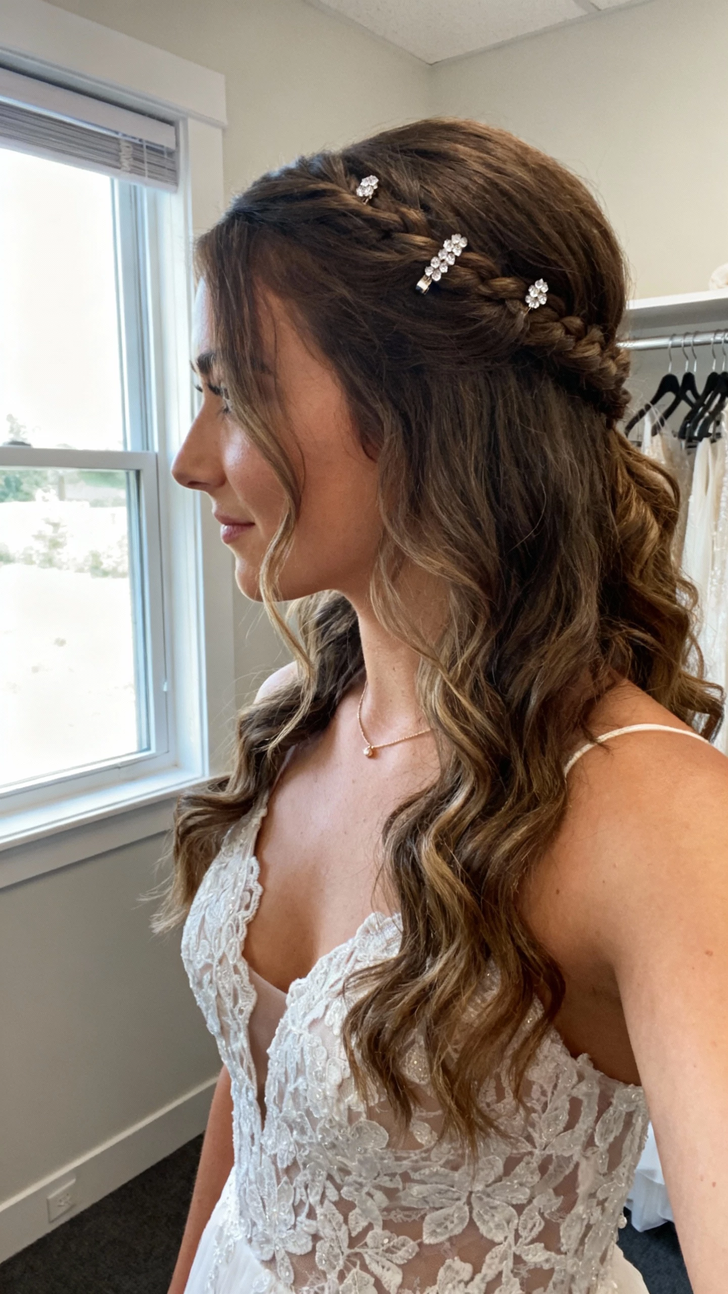 iPhone side-angle selfie cropped at neck showing a woman with half-up half-down loose curls and a hidden braid detail, lace bodice gown, small crystal hairpins, and a simple necklace; face not visible, bridal suite by a window, soft morning light, natural iPhone photo.