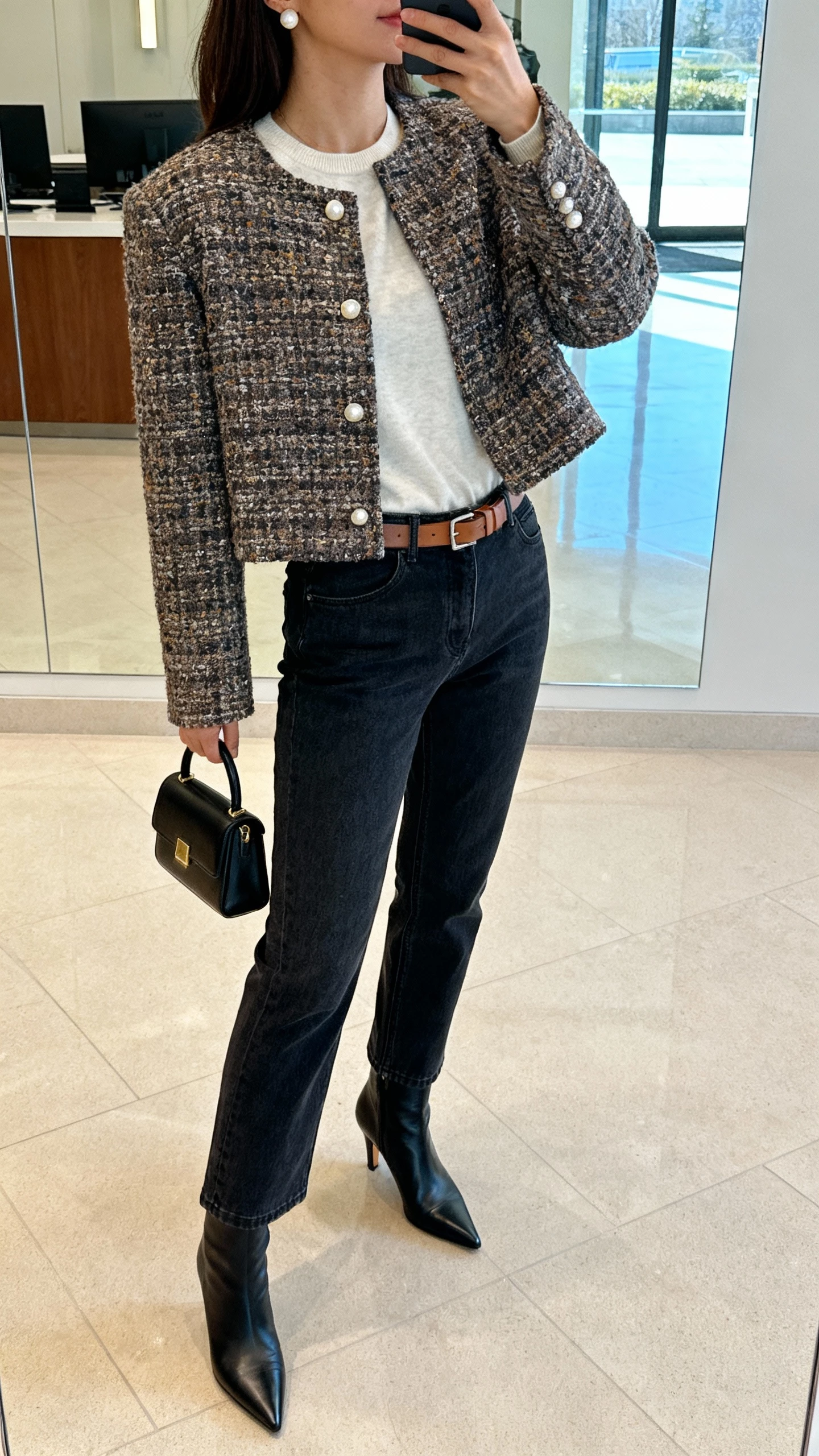 iPhone side-angle selfie cropped at neck showing a woman wearing a boxy tweed jacket, straight‑leg dark‑wash jeans, soft cashmere tee, pointed kitten‑heel boots, mini top‑handle bag, pearl studs and a slim leather belt; office lobby mirror, face not visible, natural window light, iPhone photo quality.