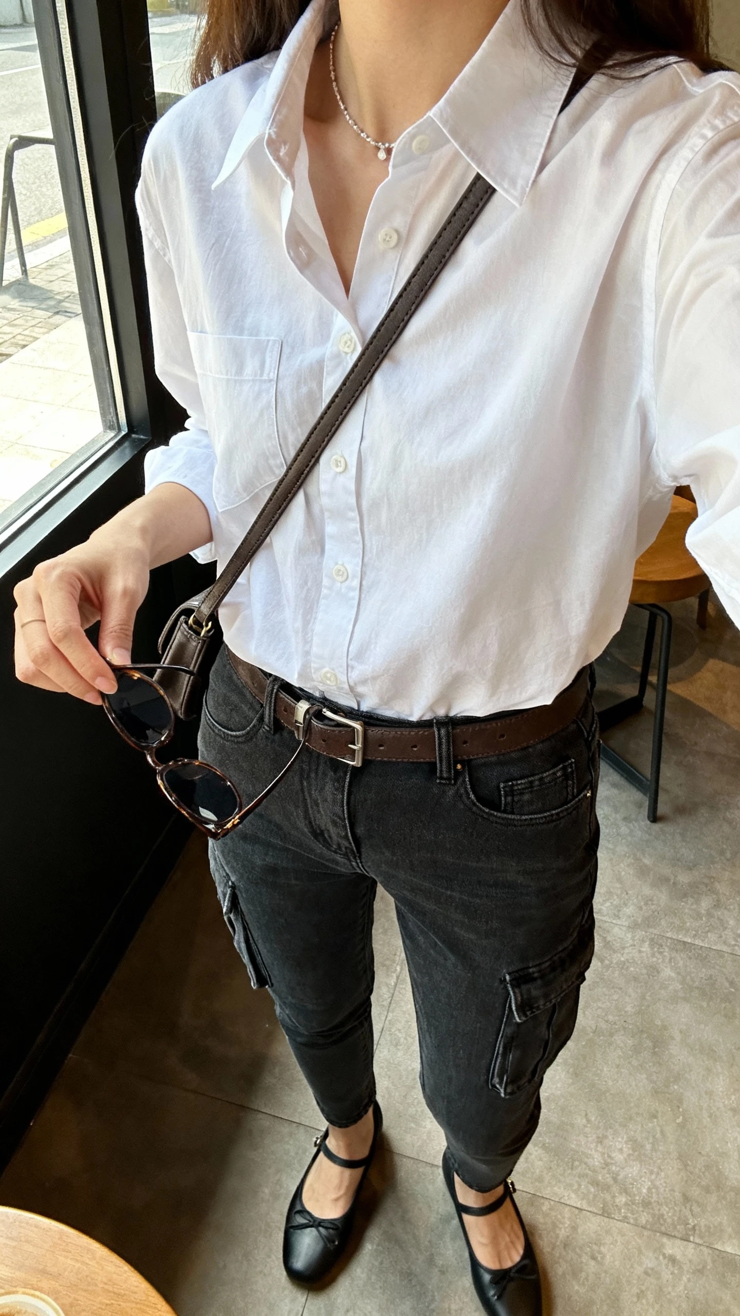 iPhone side-angle selfie cropped at shoulders showing a woman in a classic white button-down tucked into dark-wash slim cargo jeans, leather belt matched to ballet flats, structured crossbody, delicate necklace and sunglasses in hand, face not visible, coffee shop window light, natural iPhone photo.