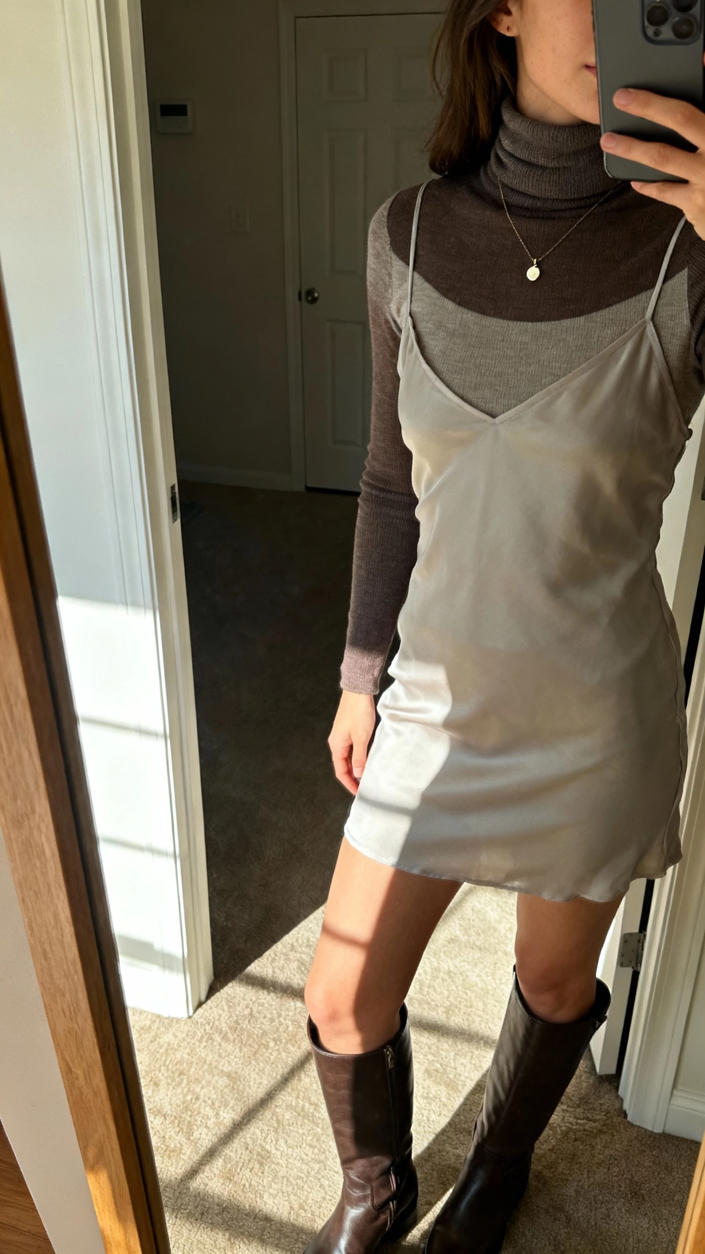 iPhone side-angle selfie cropped at shoulders showing a woman wearing a turtleneck layered under a slip dress with tall boots and a small pendant necklace, face not visible, apartment entry mirror with morning light, natural iPhone photo quality.
