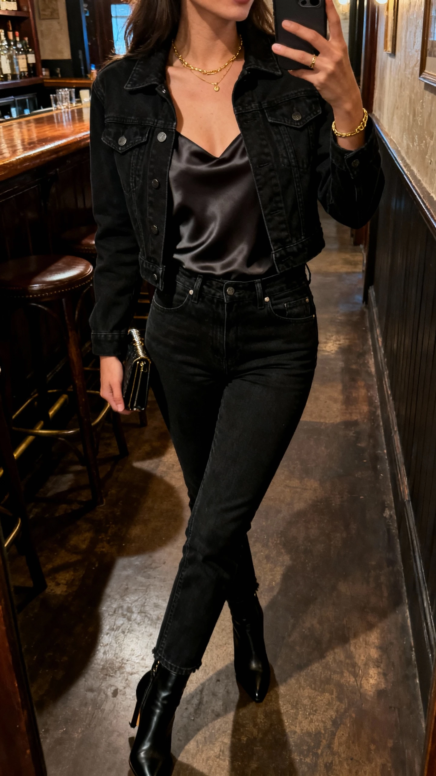 iPhone side-angle selfie of a woman in a fitted black denim jacket over a silky camisole, high-waisted black denim jeans, heeled ankle boots, delicate gold jewelry, small clutch, face not visible, dim bar hallway lighting, natural iPhone photo quality