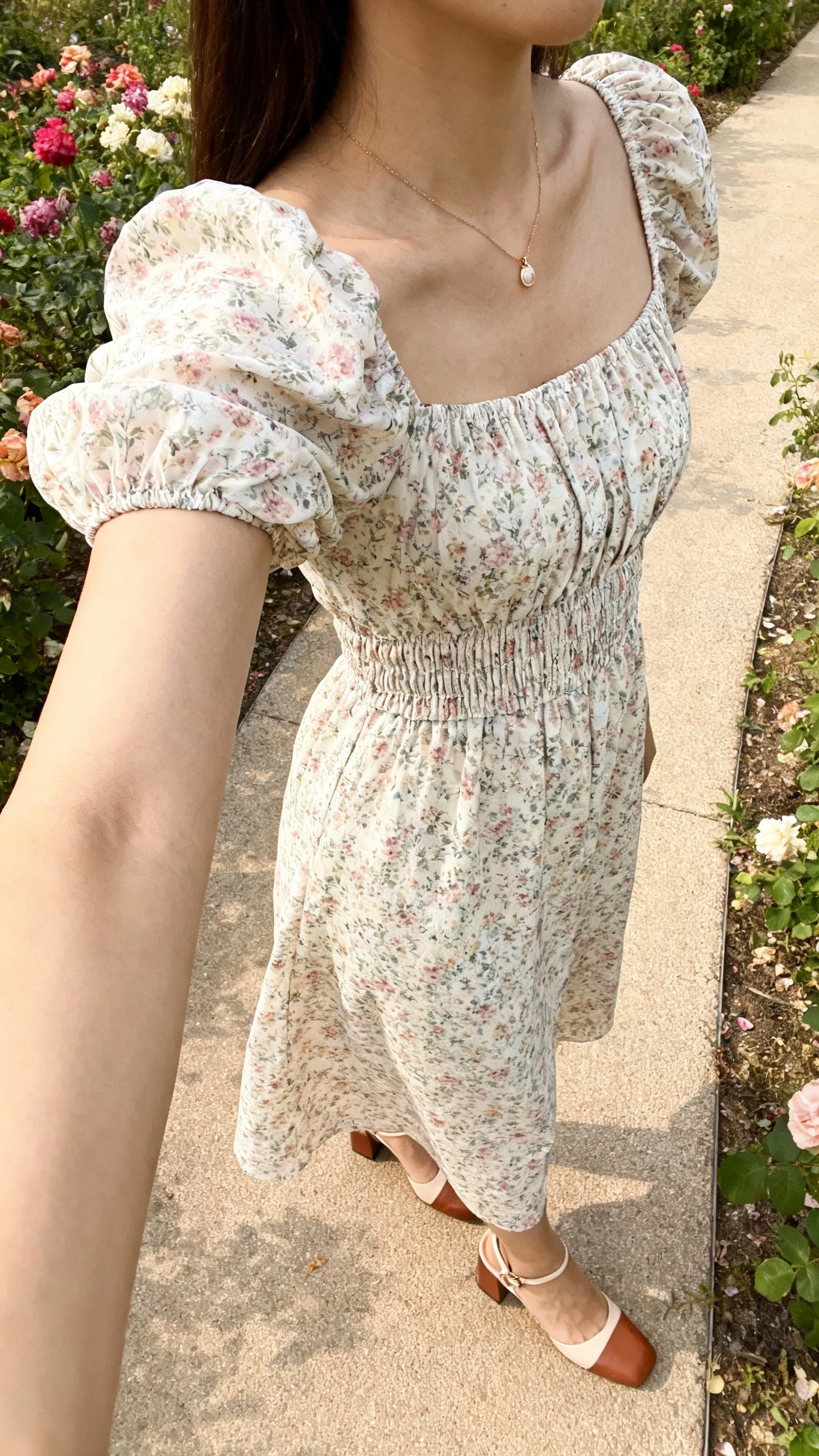 iPhone side-angle selfie of a woman in a floral puff-sleeve midi dress with a smocked waist, dainty pendant necklace, block-heel sandals, face not visible, taken in a garden path near blooming flowers, soft afternoon daylight, natural iPhone aesthetic.