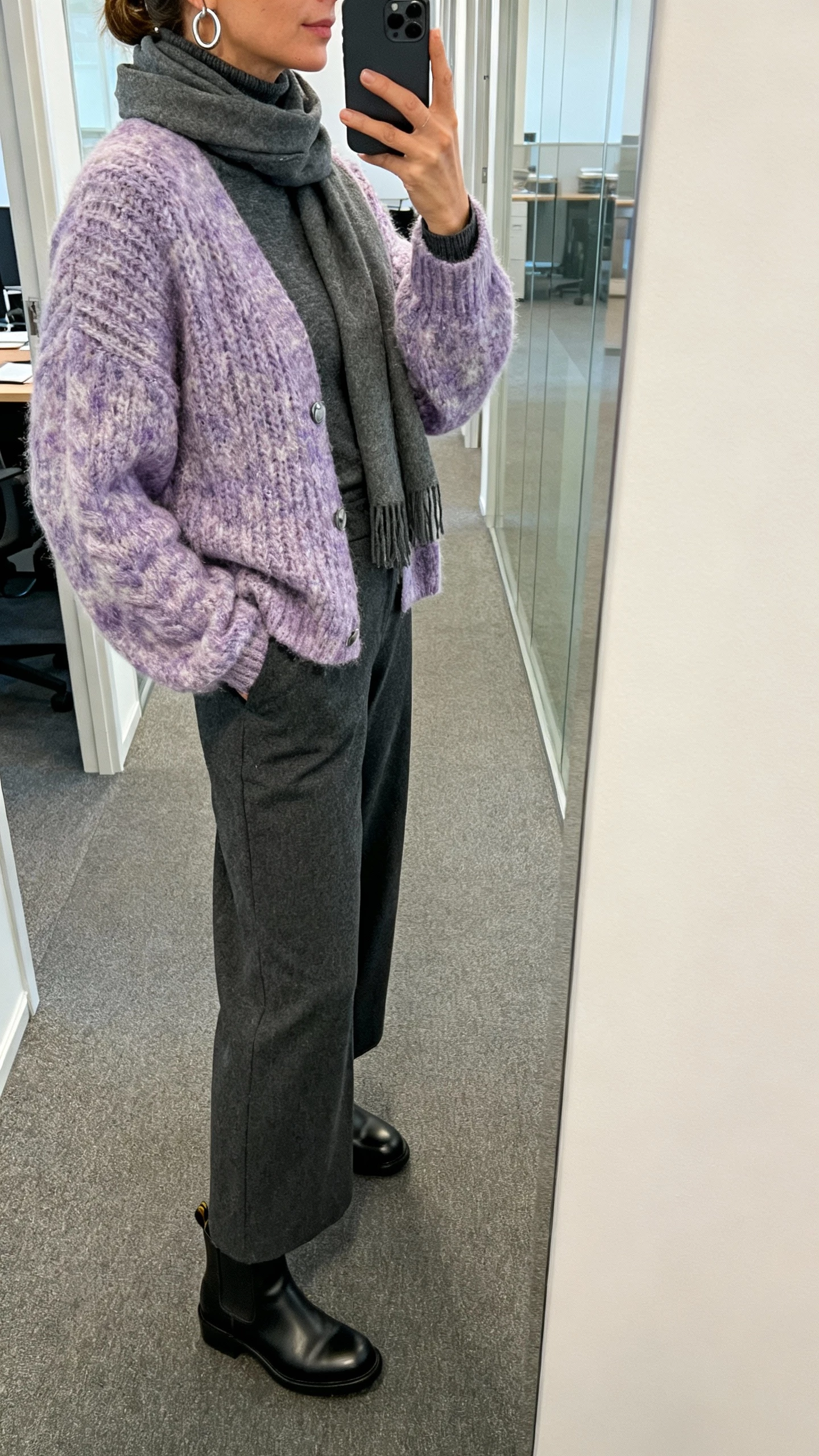 iPhone side-angle selfie of a woman in a frosted lilac chunky cardigan over a charcoal turtleneck, charcoal wide-leg trousers, black chelsea boots, silver hoop earrings, slate scarf, face not visible, office hallway mirror with soft indoor light, natural iPhone aesthetic