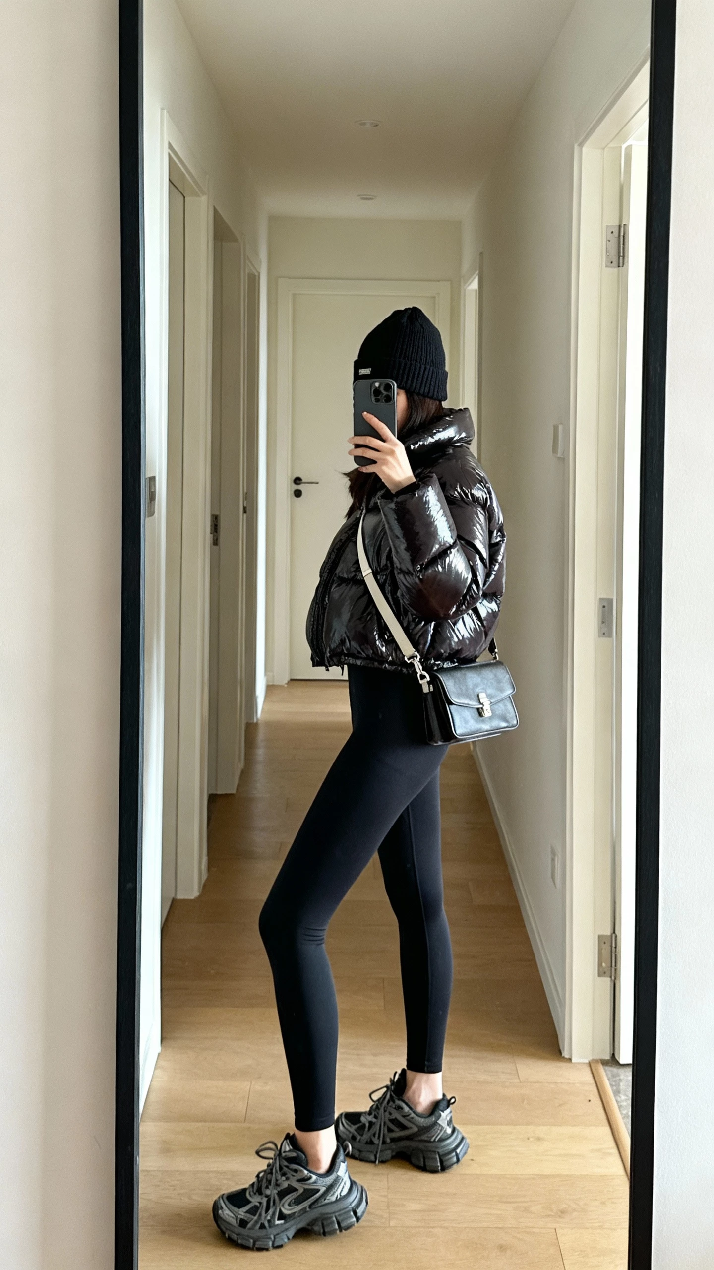 iPhone side-angle selfie of a woman in a glossy puffer jacket, sleek black leggings, chunky sneakers, beanie, crossbody bag, face not visible, apartment hallway mirror, natural daylight, casual iPhone aesthetic.