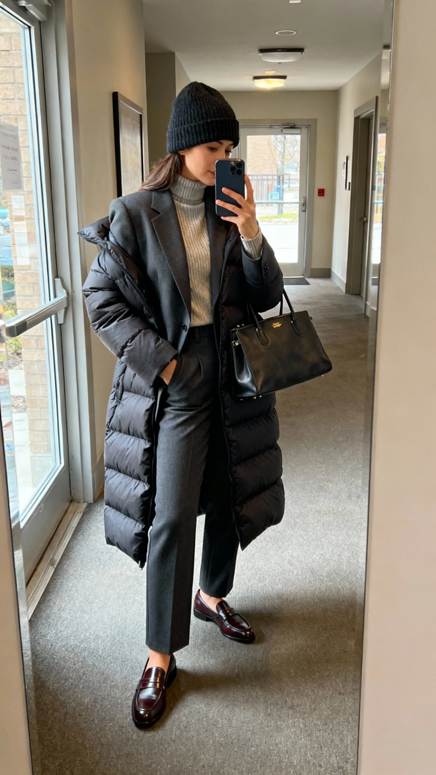 iPhone side-angle selfie of a woman in a longline puffer layered over a tailored blazer and fine-knit turtleneck, straight-leg trousers, sleek leather loafers, structured tote, wool beanie, face not visible, hallway mirror in an apartment lobby, natural daylight from glass door, casual iPhone photo