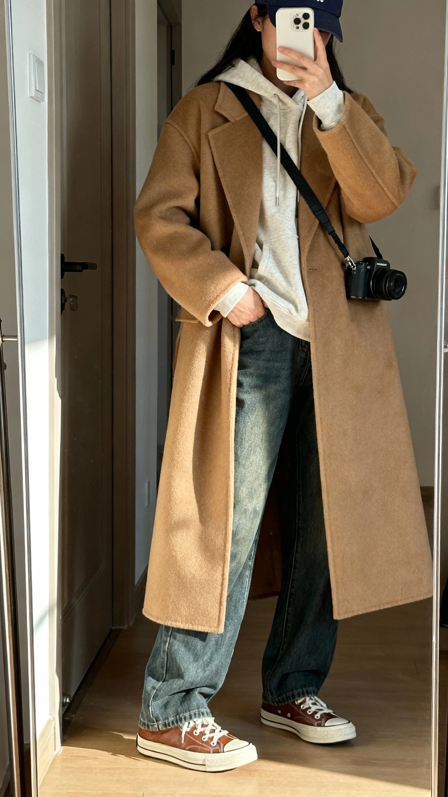 iPhone side-angle selfie of a woman in a longline camel wool coat layered over a heavyweight cotton hoodie, straight-leg jeans, retro leather sneakers, crossbody camera bag, and a baseball cap; face not visible, hallway mirror near the door, morning light, casual iPhone photo.