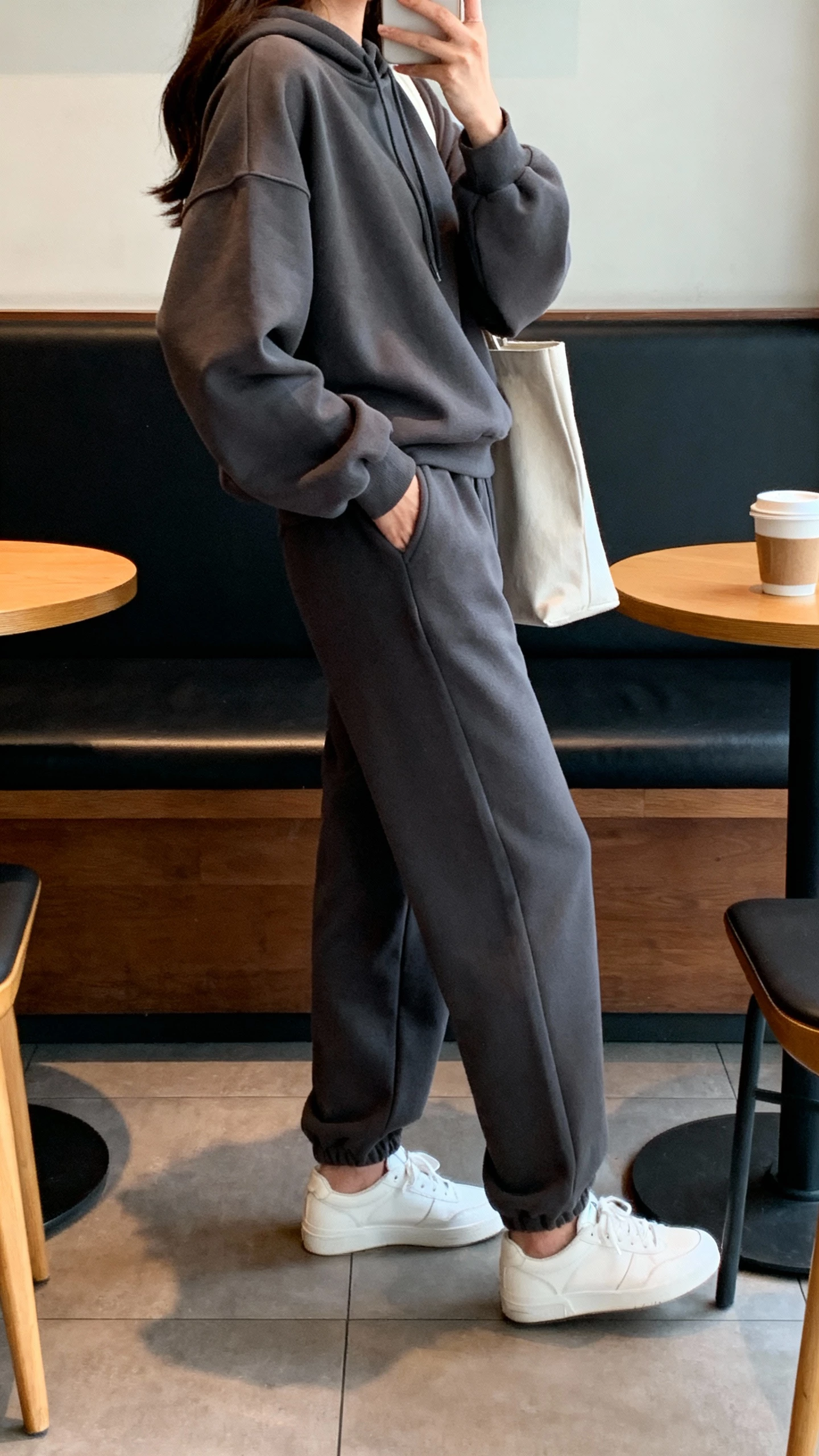 iPhone side-angle selfie of a woman in a monochrome charcoal tracksuit (relaxed hoodie and joggers), sleek white trainers, minimalist tote, face not visible, coffee shop booth, indoor natural light, casual iPhone photo