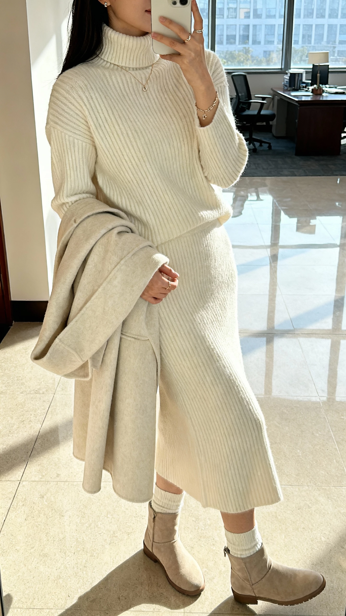 iPhone side-angle selfie of a woman in a monochrome knit-on-knit set: ribbed turtleneck sweater with matching knit skirt, tonal coat over arm, sock boots, and delicate jewelry, face not visible, office lobby window light, natural iPhone quality.