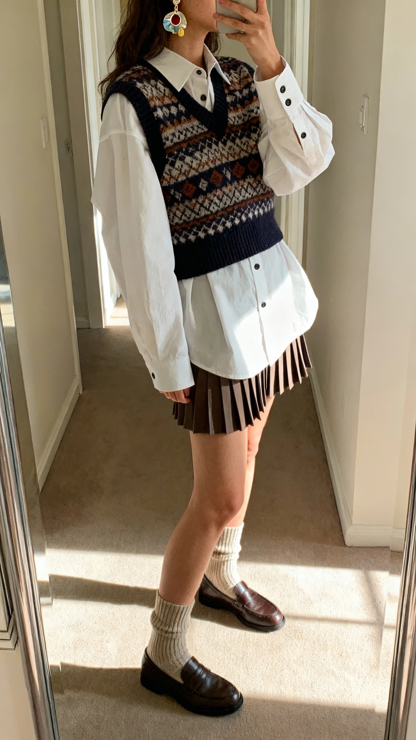 iPhone side-angle selfie of a woman in a patterned sweater vest layered over an oversized white button-up, pleated mini skirt, ribbed socks, loafers, and quirky artsy earrings, face not visible, hallway mirror with morning light, casual iPhone aesthetic.