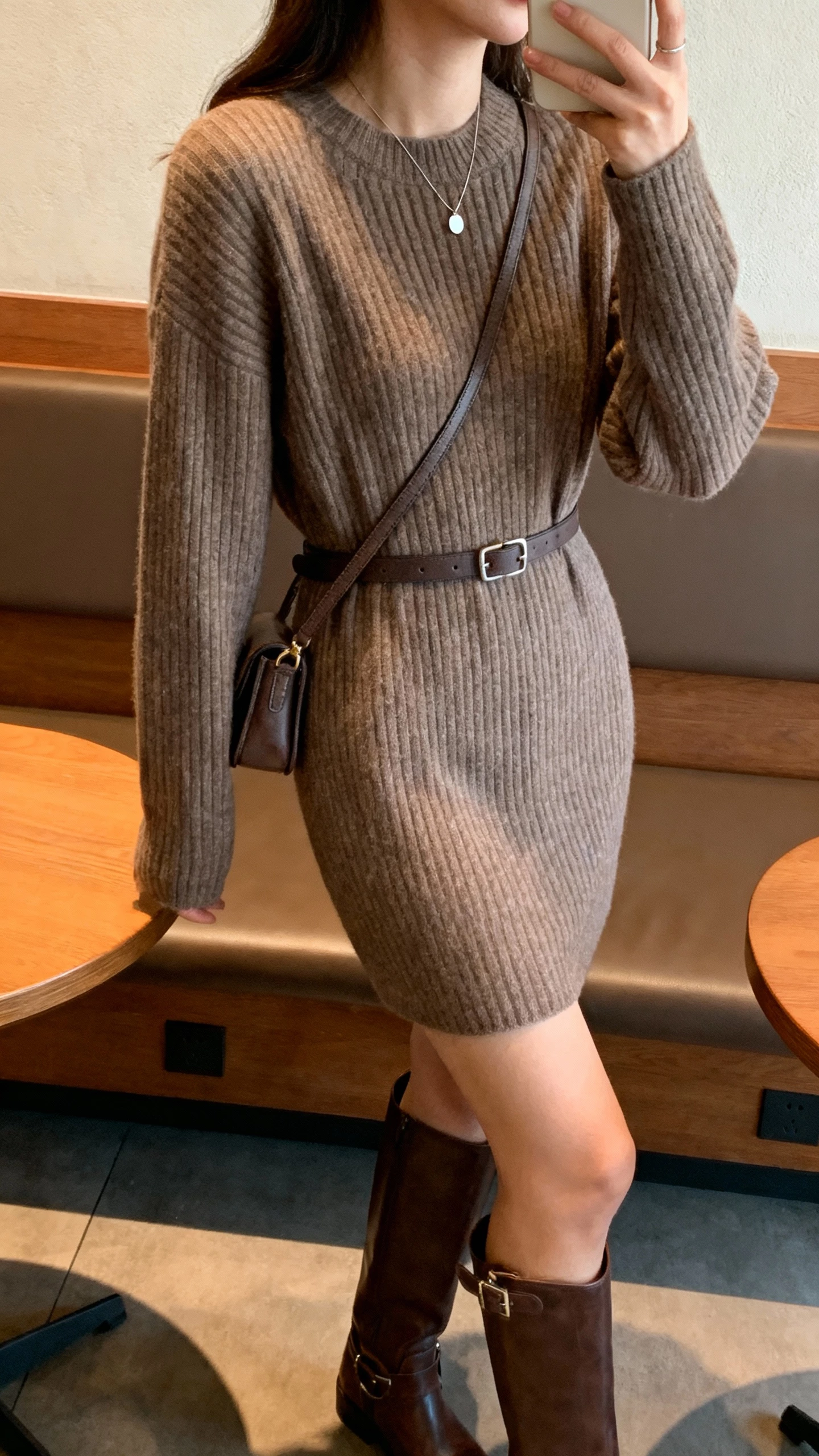 iPhone side-angle selfie of a woman in a ribbed sweater dress with a waist belt, knee-high leather boots, minimal pendant necklace, small crossbody bag, face not visible, coffee shop booth, warm indoor lighting, natural iPhone aesthetic.
