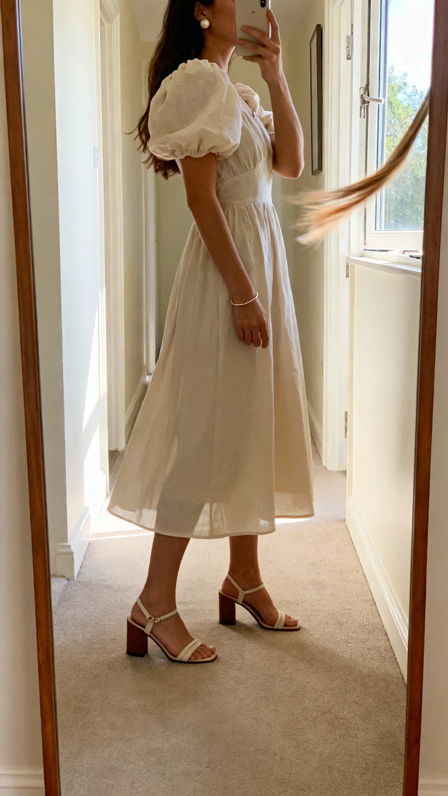 iPhone side-angle selfie of a woman in a romantic puff-sleeve midi dress, block-heel sandals, pearl studs, dainty bracelet, face not visible, hallway mirror catching a soft breeze from an open window, morning light, natural iPhone aesthetic.