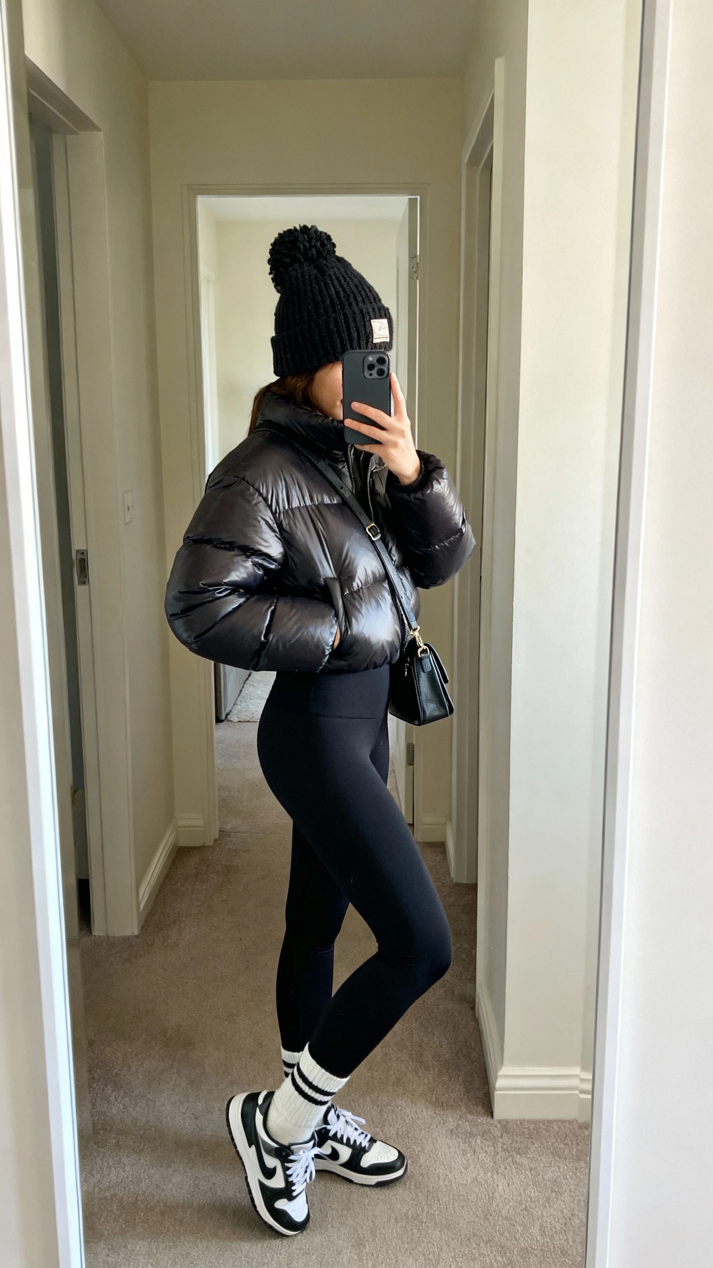 iPhone side-angle selfie of a woman in a sleek puffer jacket, black high-waist leggings, chunky knit beanie, sock sneakers, crossbody bag, face not visible, hallway mirror, soft indoor lighting, natural iPhone photo quality