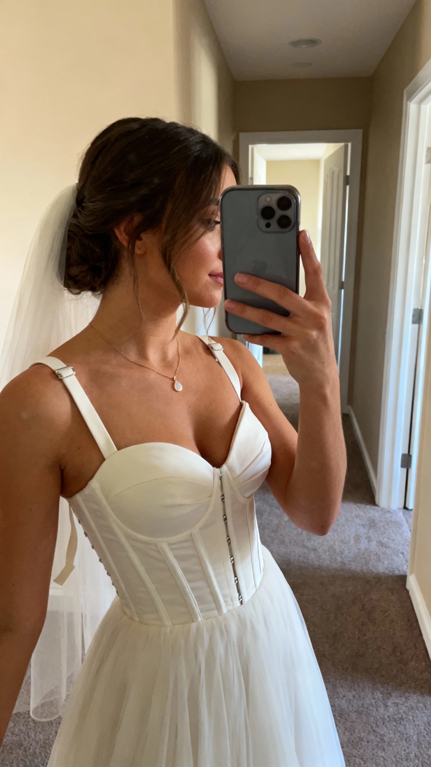 iPhone side-angle selfie of a woman in a sweetheart-neckline wedding dress with built-in cups, boned bodice, and secure straps for lift and shape; delicate pendant and fingertip veil, face not visible, hallway mirror, warm indoor lighting, natural iPhone photo quality