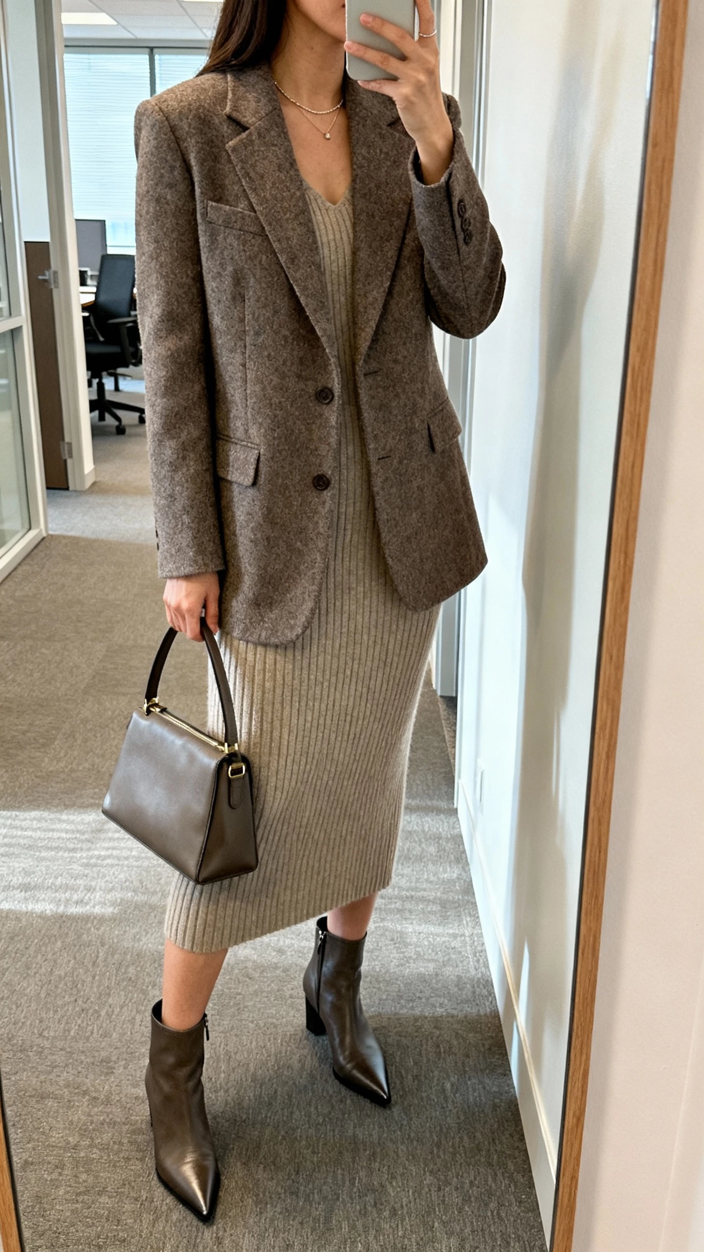 iPhone side-angle selfie of a woman in a tailored wool blazer over a ribbed knit dress, pointed ankle boots, structured top-handle bag, minimal necklace, face not visible, office hallway mirror, morning light, natural iPhone aesthetic.