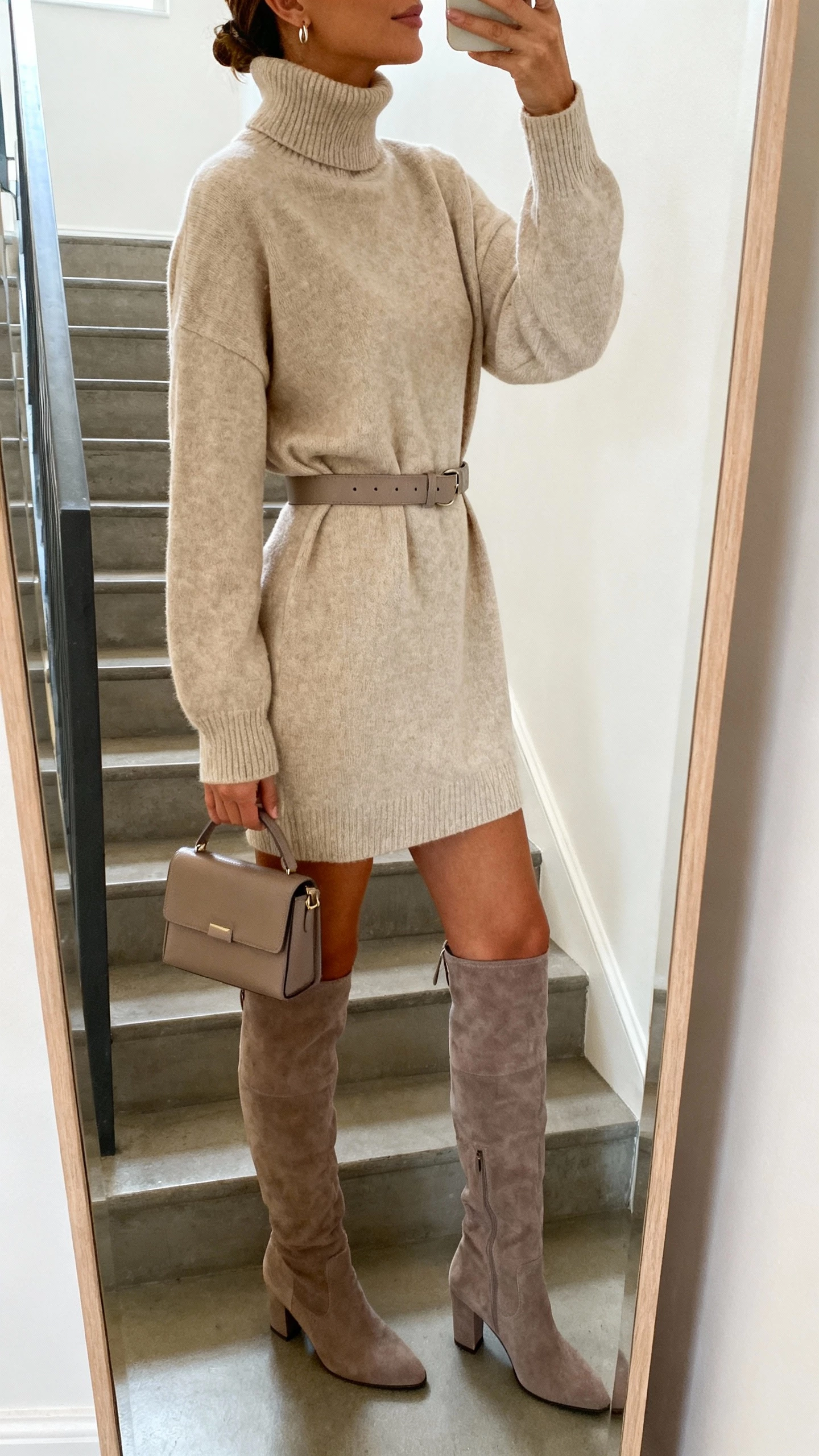 iPhone side-angle selfie of a woman in a tonal beige turtleneck sweater dress with a waist belt, taupe over-the-knee boots, structured mini bag, subtle earrings, face not visible, stairwell mirror, soft daylight, natural iPhone photo quality.