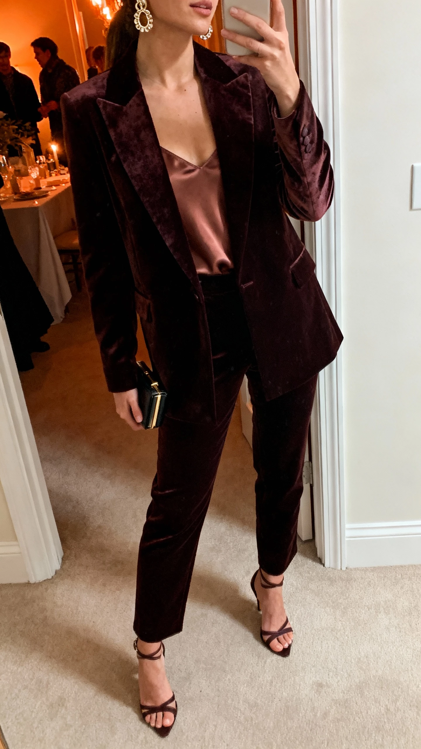 iPhone side-angle selfie of a woman in a velvet blazer and matching tailored trousers with a satin cami, strappy heels, statement earrings, and a tiny clutch, face not visible, hallway outside a party with warm indoor lighting, natural iPhone photo quality.