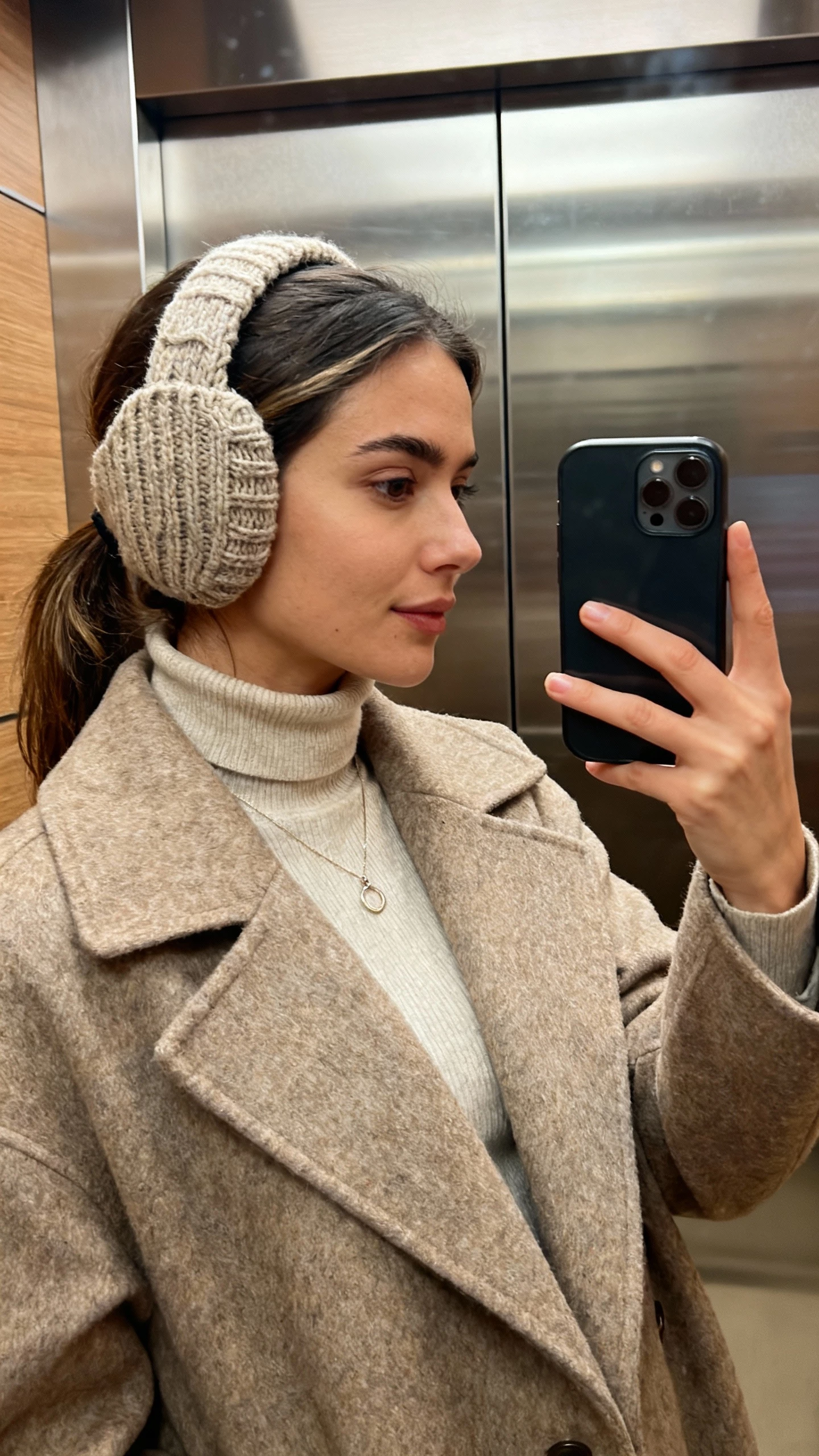 iPhone side-angle selfie of a woman in an elevated knit ear-warmer headband that shows her hair, turtleneck, wool overcoat, delicate pendant, face not visible, elevator mirror, warm indoor lighting, natural iPhone photo quality.