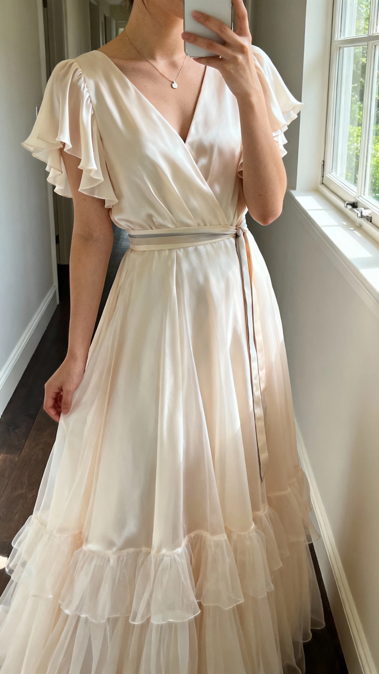 iPhone side-angle selfie of a woman in an ethereal silk chiffon bridal dress with airy layered skirt, soft flutter sleeves, thin sash at waist, minimalist pendant, face not visible, hallway near window light, natural iPhone photo quality