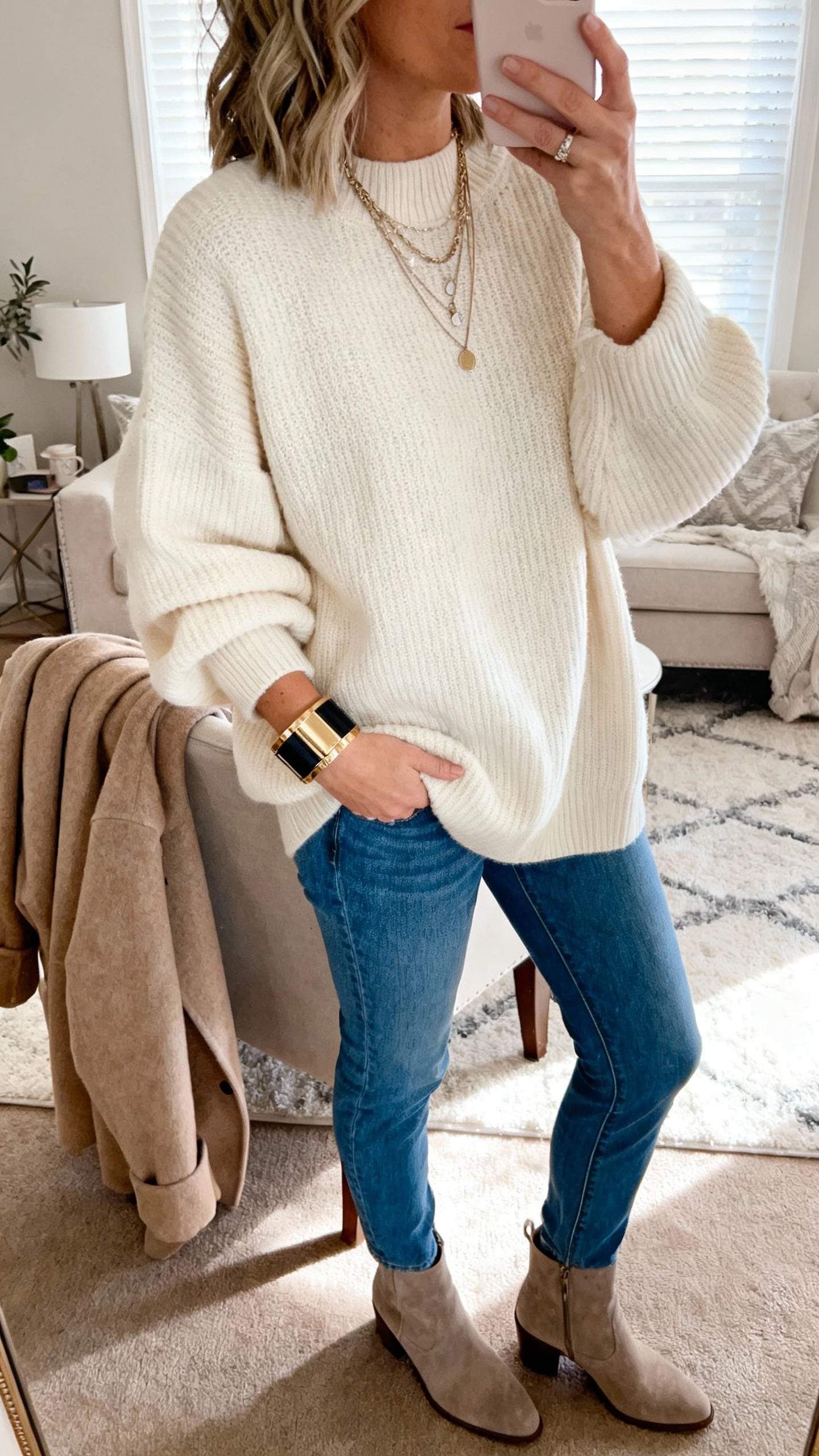 iPhone side-angle selfie of a woman in an oversized cream knit sweater, blue straight jeans, ankle boots, layered necklaces, bold cuff bracelet, wool coat over chair; cozy living room, face not visible, afternoon window light, natural iPhone photo quality.