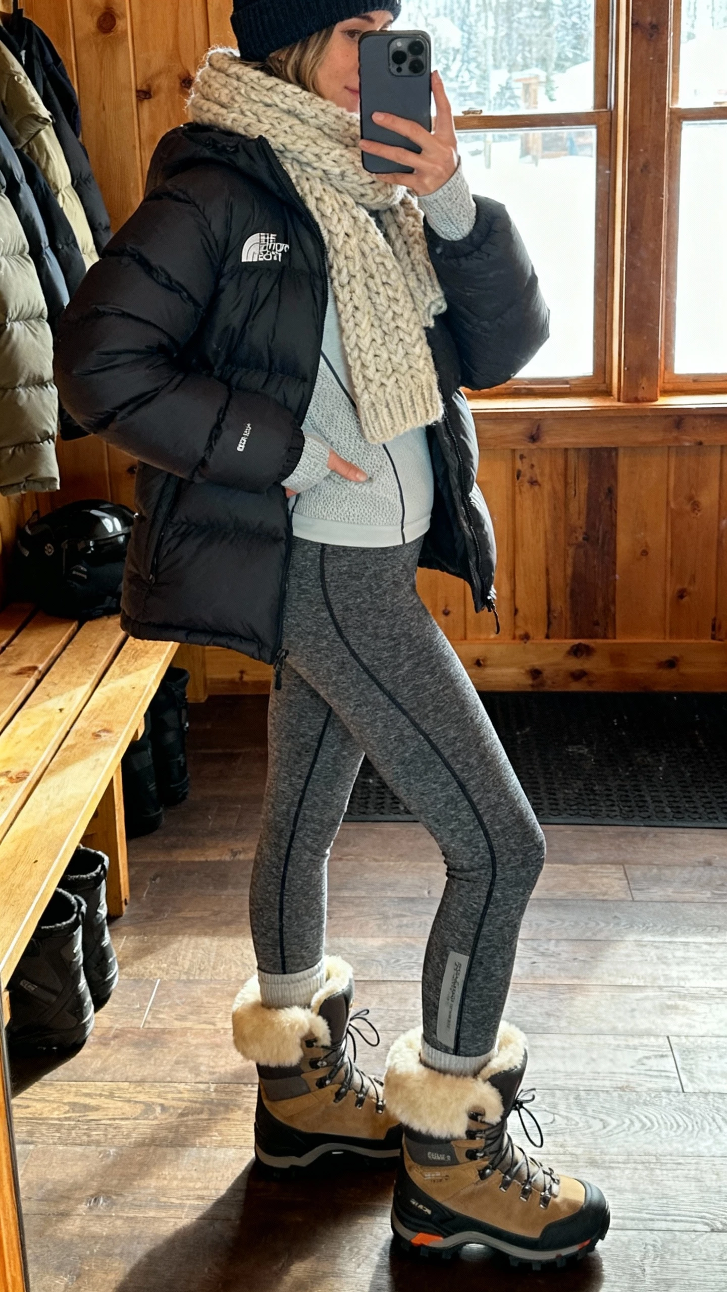 iPhone side-angle selfie of a woman in shearling-lined hiker boots, puffer jacket, thermal sweater, fleece-lined leggings, chunky knit scarf, and beanie, face not visible, ski-lodge mudroom setting, afternoon window light, casual iPhone aesthetic.