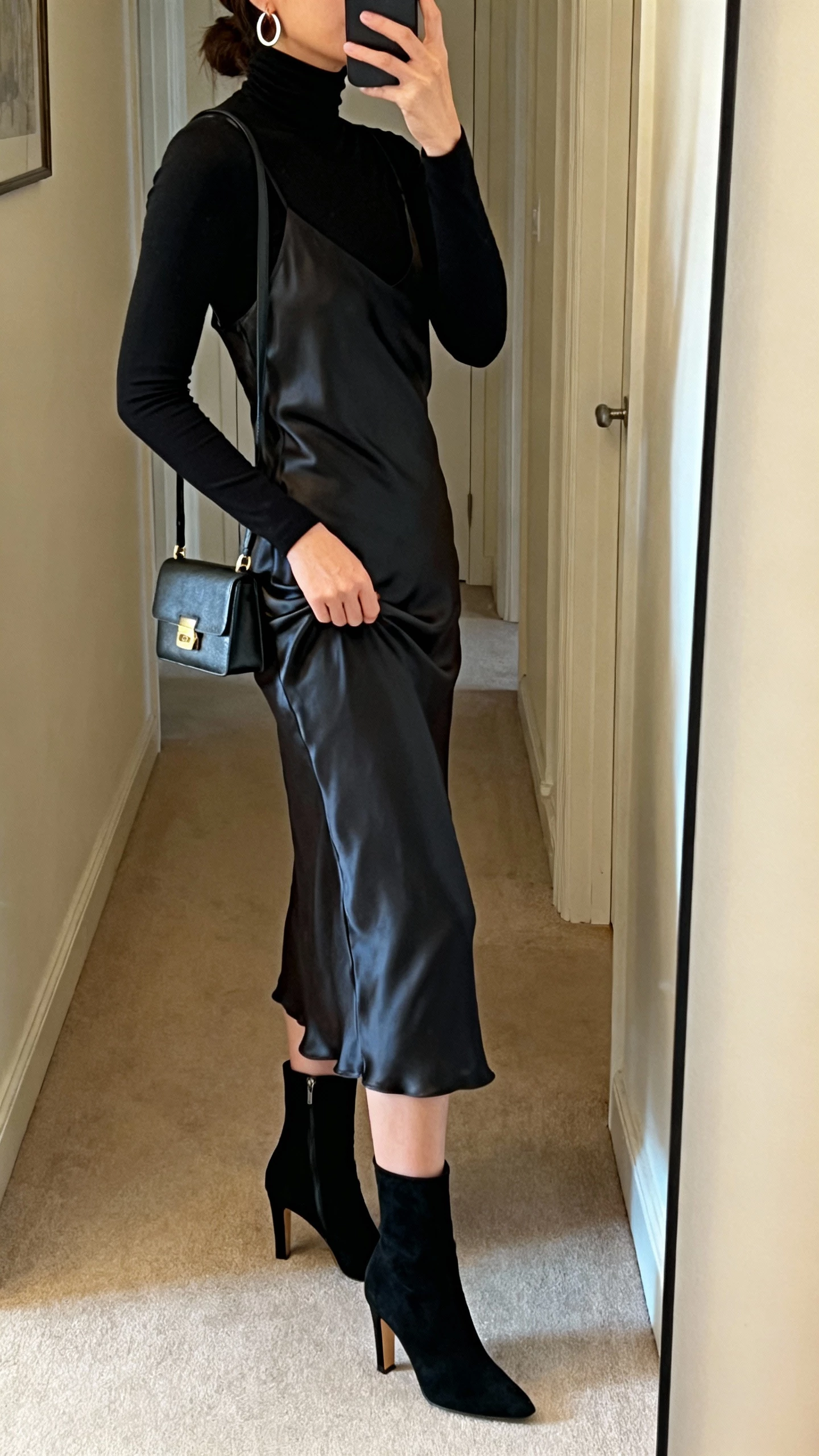 iPhone side-angle selfie of a woman layering a black fitted turtleneck under a satin slip dress, heeled booties, small shoulder bag, hoop earrings, face not visible, hallway mirror, warm indoor lighting, natural iPhone photo quality.