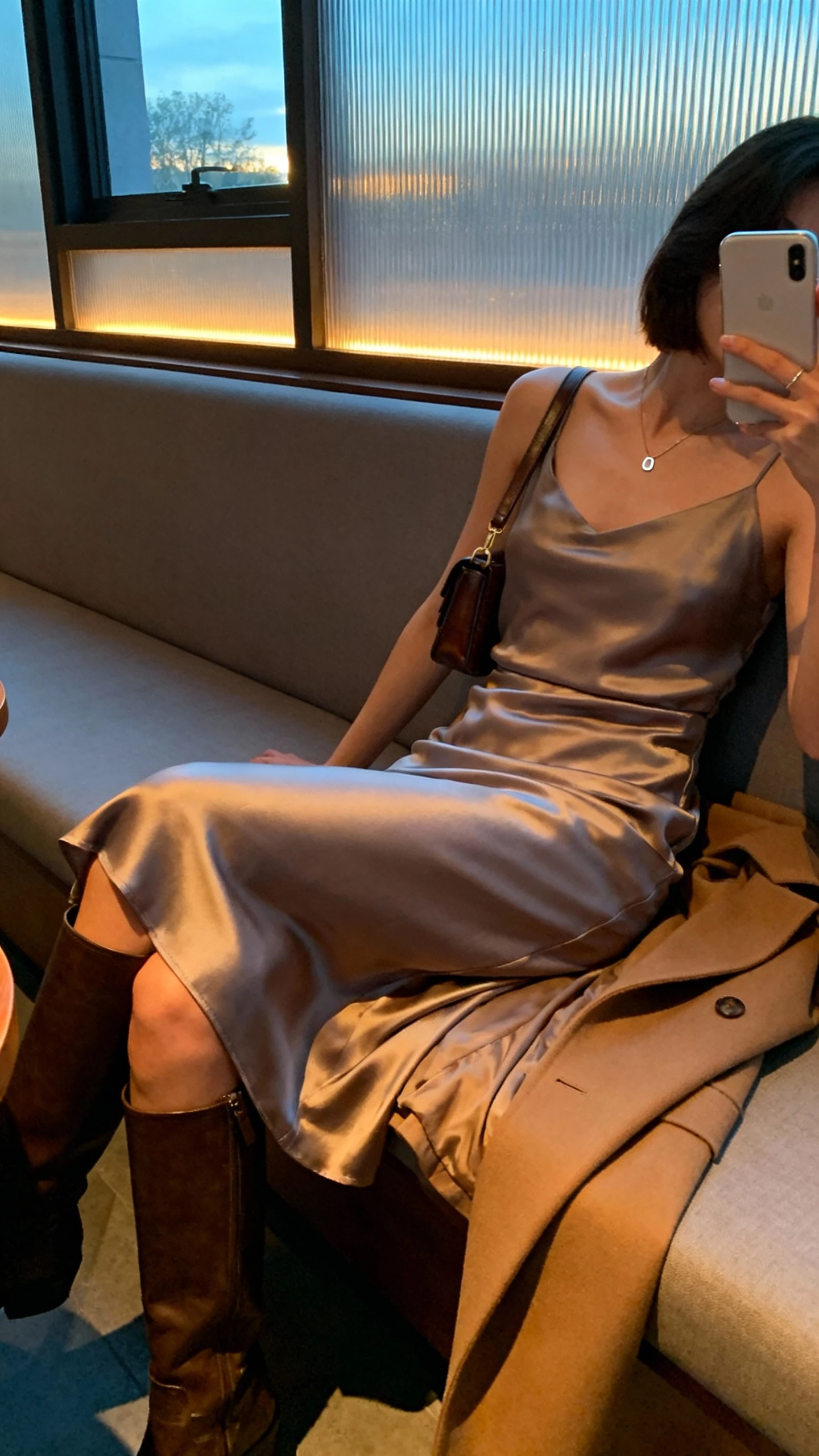 iPhone side-angle selfie of a woman seated at a lounge banquette wearing a satin midi dress with knee-high boots, minimal pendant necklace, small shoulder bag, and a longline coat draped over the seat, face not visible, evening indoor lighting with window glow, natural iPhone aesthetic.