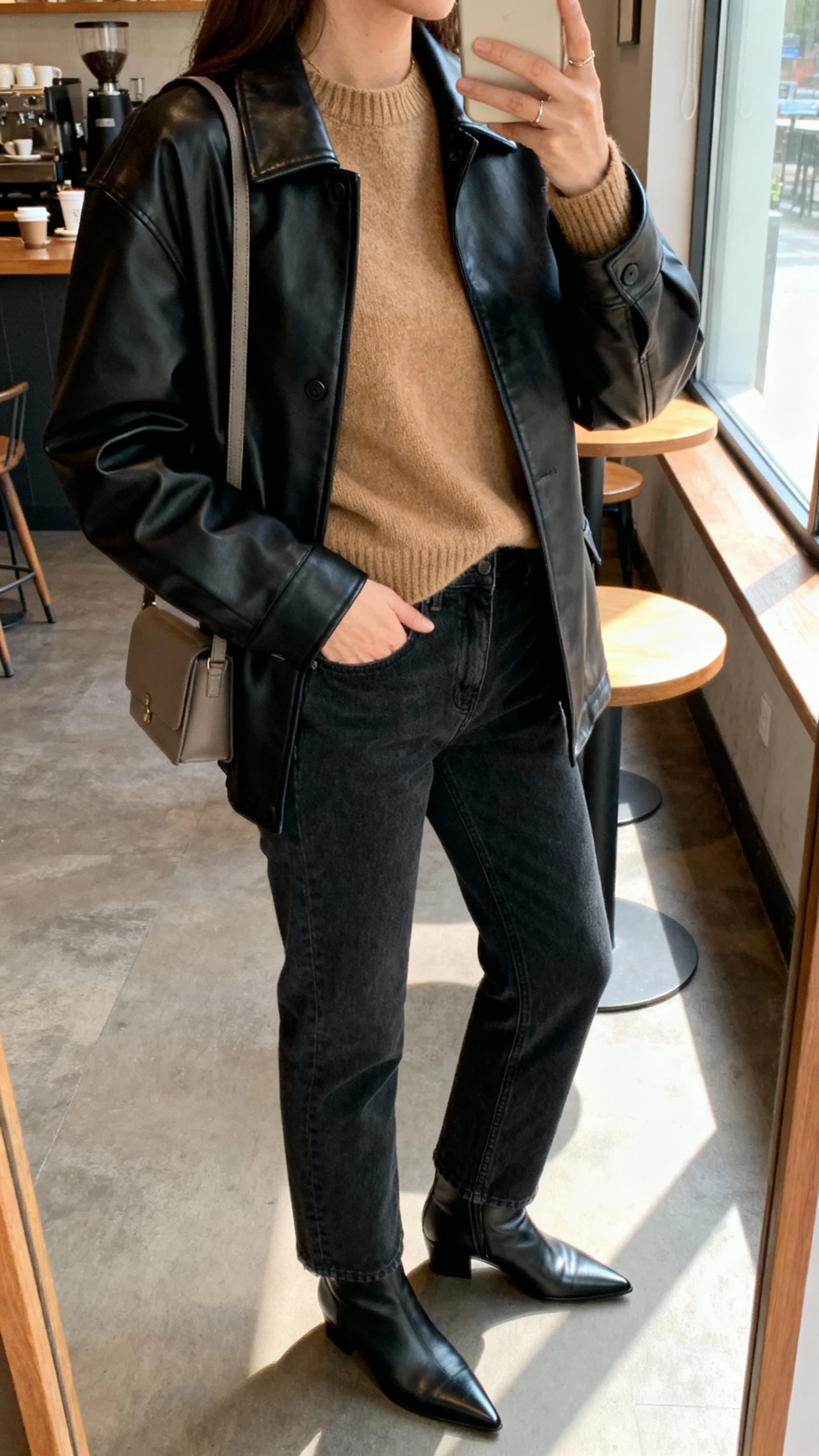 iPhone side-angle selfie of a woman wearing a black leather jacket over a camel cashmere sweater, straight dark jeans, pointed boots, and a minimal shoulder bag; face not visible, taken in a coffee shop near window light, casual iPhone aesthetic.