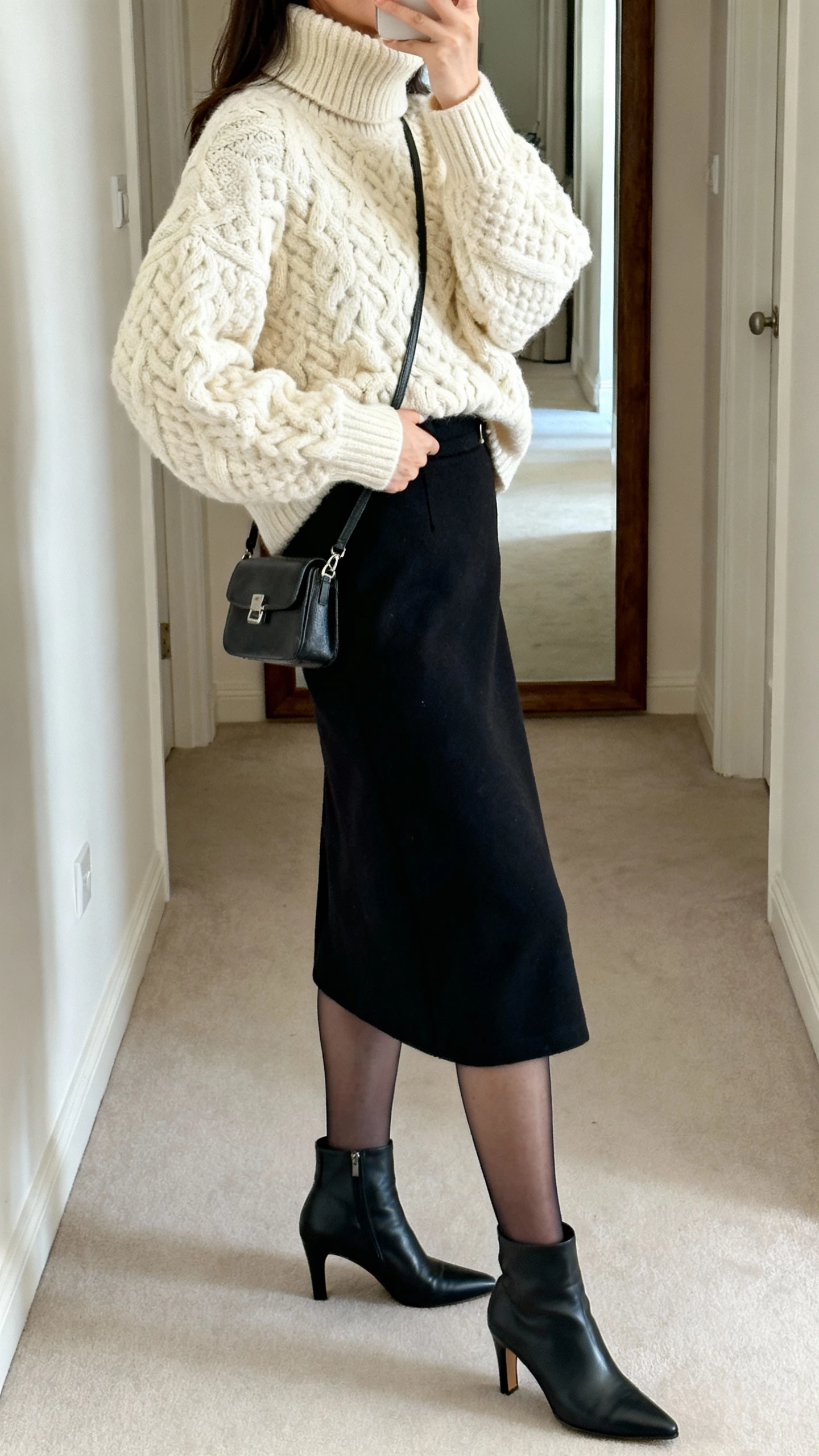 iPhone side-angle selfie of a woman wearing a chunky cream turtleneck sweater tucked into a black midi skirt, sheer tights, heeled ankle boots, small crossbody bag, face not visible, hallway mirror, soft indoor lighting, natural iPhone photo.
