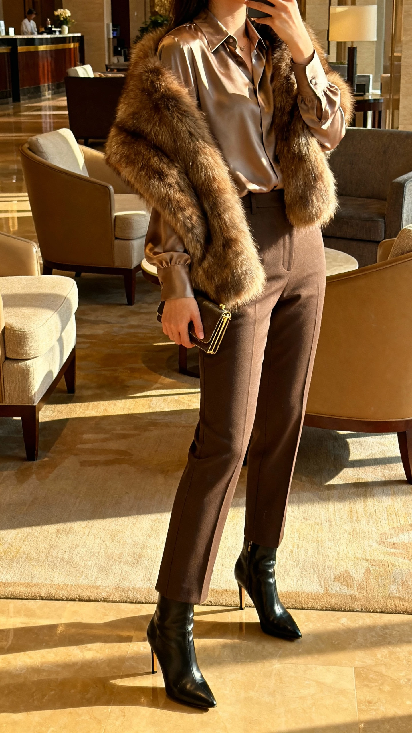 iPhone side-angle selfie of a woman wearing a faux-fur stole over a satin blouse, tailored trousers, and heeled boots, small clutch in hand, face not visible, hotel lobby seating area with warm indoor lighting, natural iPhone photo quality.