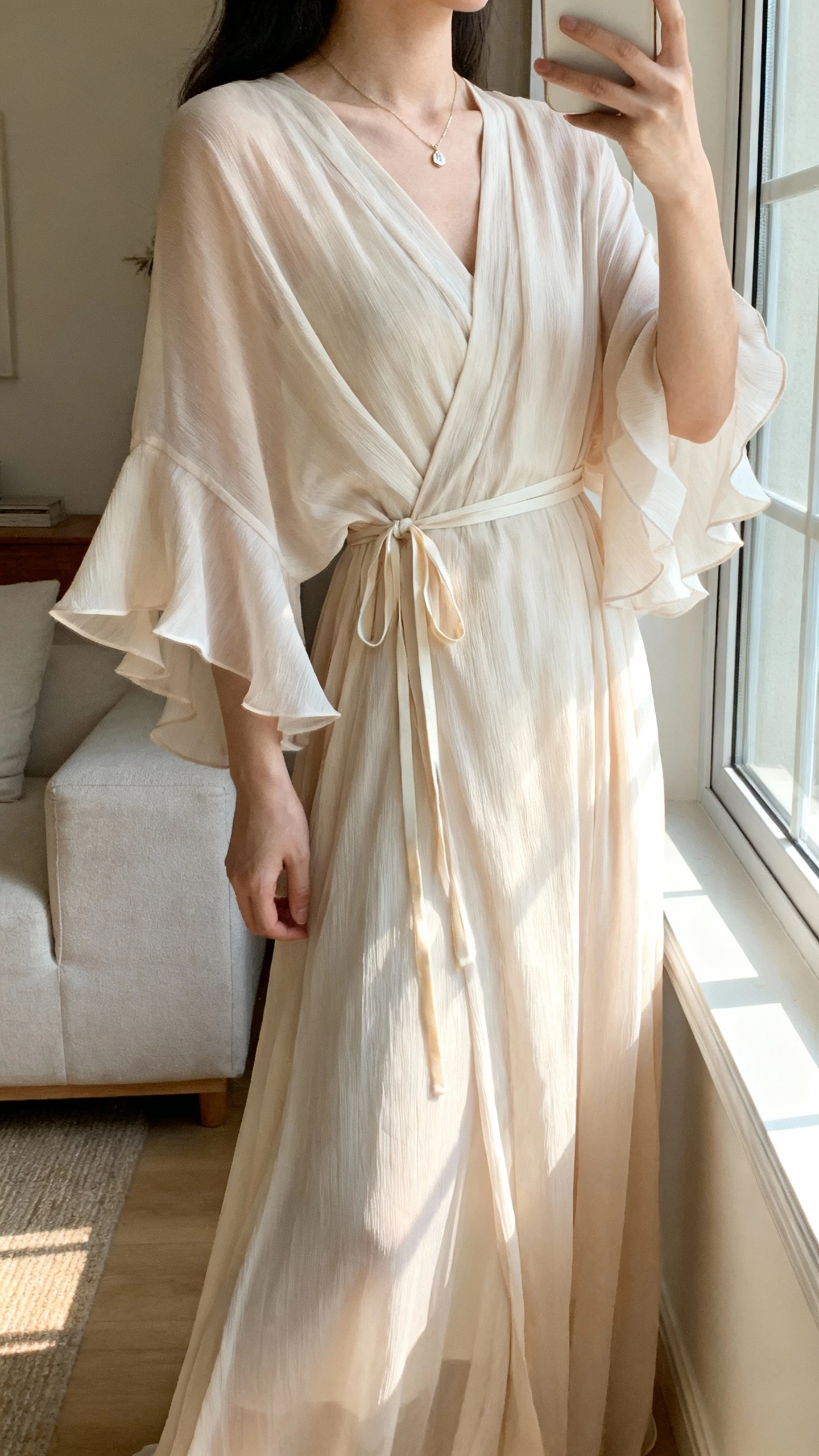iPhone side-angle selfie of a woman wearing a flowing chiffon boho wrap gown with flutter sleeves, thin waist tie, dainty pendant necklace, face not visible, living room near window, soft morning light, natural iPhone photo quality