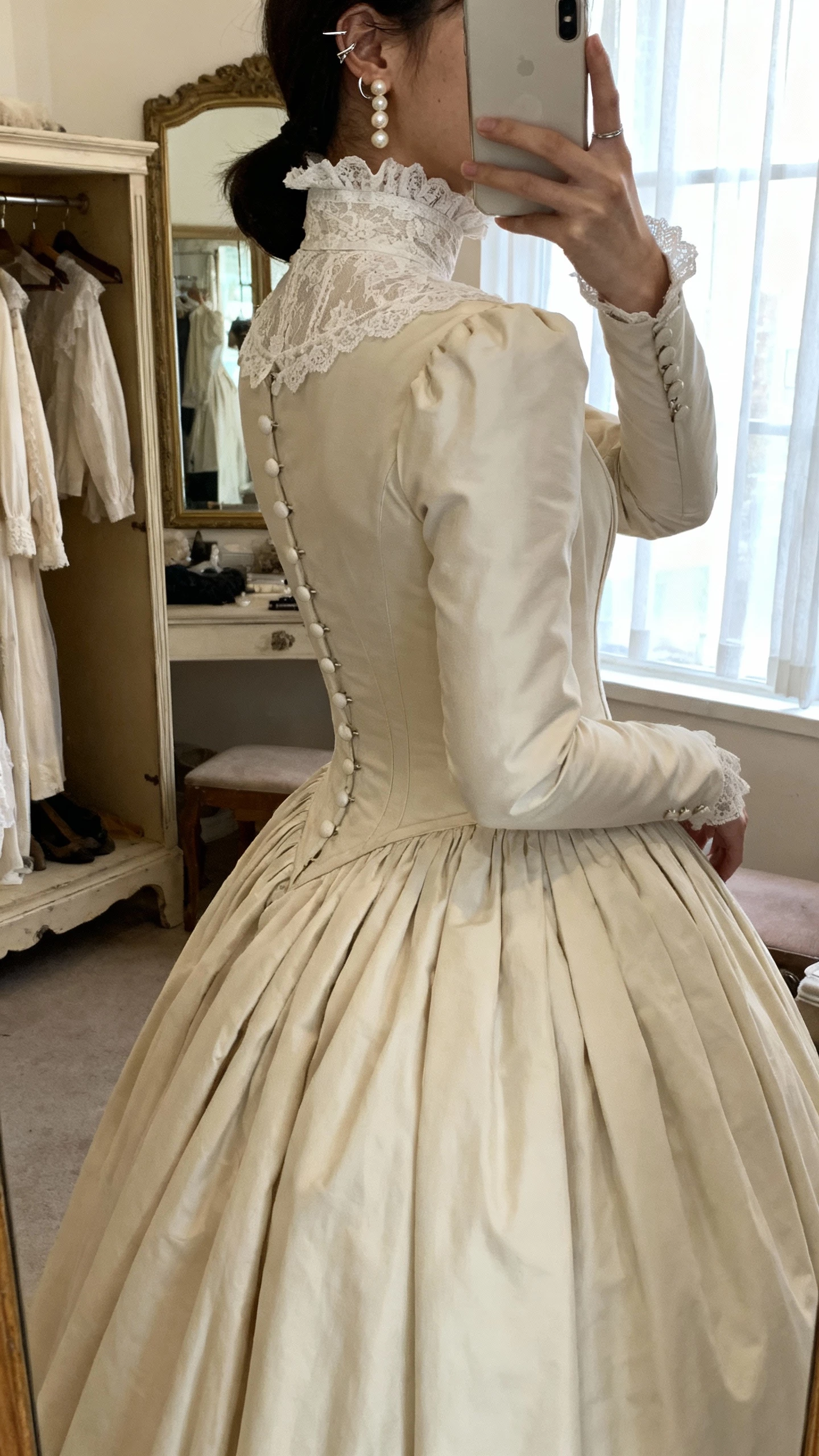 iPhone side-angle selfie of a woman wearing a high-neck Victorian-inspired ball gown with lace high collar, long fitted sleeves, button-back detail, and edgy pearl ear cuff, face not visible, vintage dressing room mirror, soft window light, natural iPhone aesthetic
