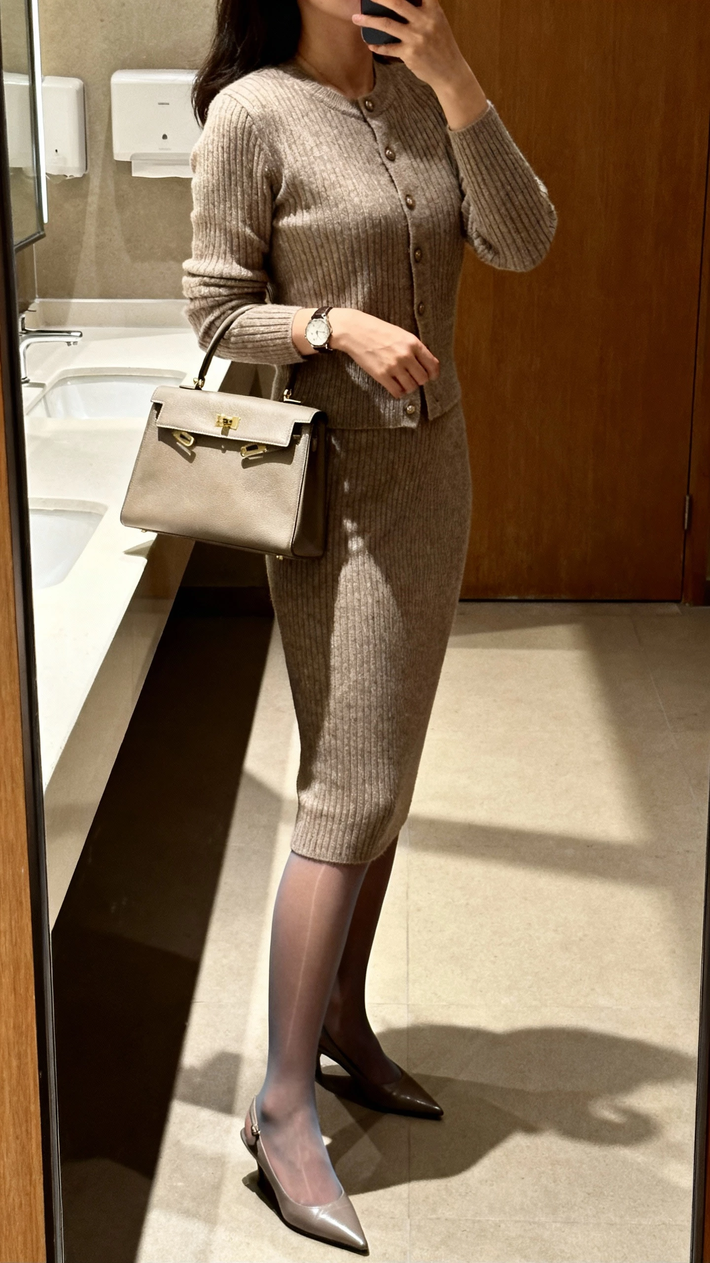 iPhone side-angle selfie of a woman wearing a knit skirt set (fitted cardigan and pencil skirt) with sheer tights, pointed slingbacks, top-handle bag, and watch, face not visible, office restroom mirror before evening plans, warm indoor lighting.
