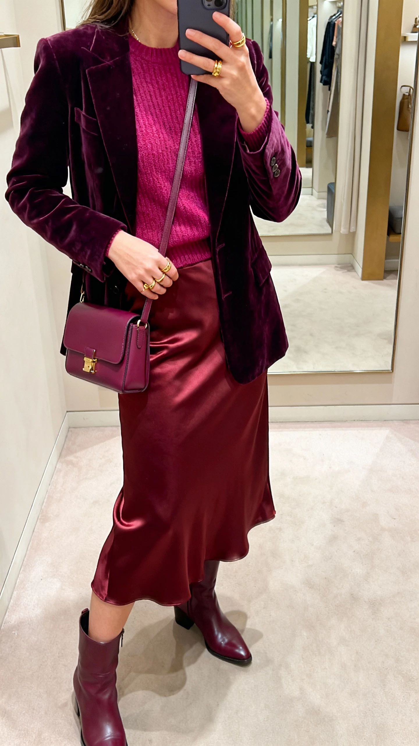 iPhone side-angle selfie of a woman wearing a monochrome berry mix: plum velvet blazer, raspberry knit top, burgundy satin midi skirt, wine leather boots, berry crossbody, gold rings, face not visible, boutique dressing room mirror, warm indoor light, iPhone quality