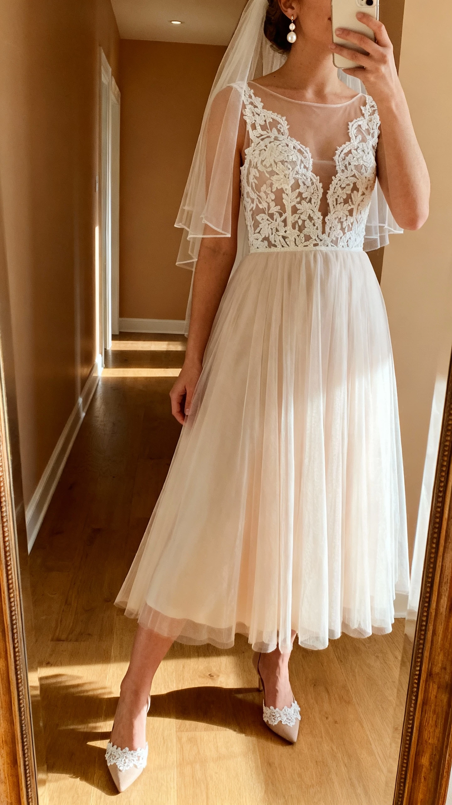 iPhone side-angle selfie of a woman wearing a romantic lace A-line wedding dress with sheer lace bodice, soft tulle skirt, fingertip veil, pearl drop earrings, and lace-trimmed heels, face not visible, hallway mirror with morning light, natural iPhone aesthetic.