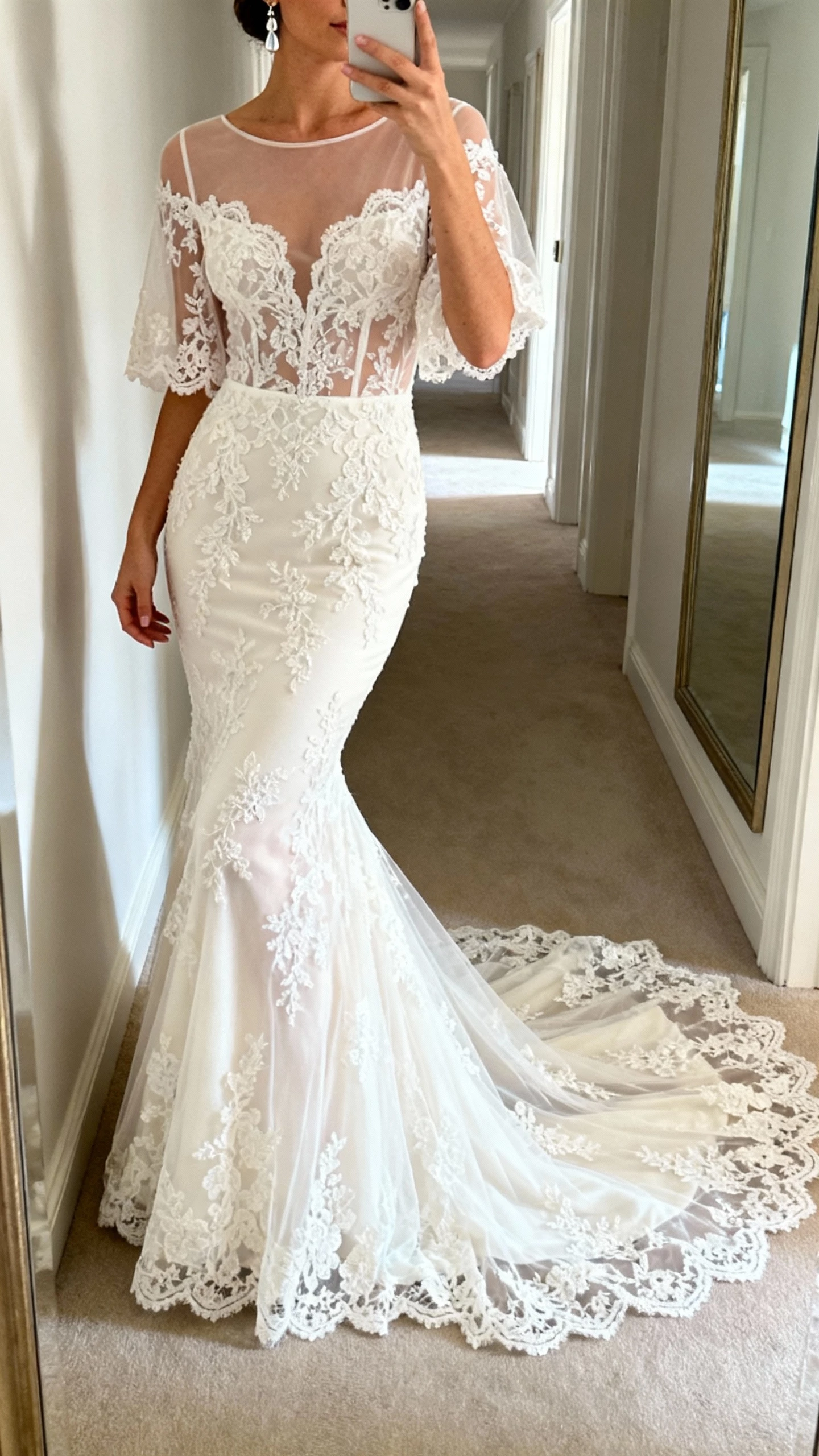 iPhone side-angle selfie of a woman wearing a romantic lace mermaid wedding dress with a sheer illusion bodice, delicate lace sleeves, and chapel-length lace train, dainty drop earrings, face not visible, hallway mirror with soft morning light, natural iPhone photo