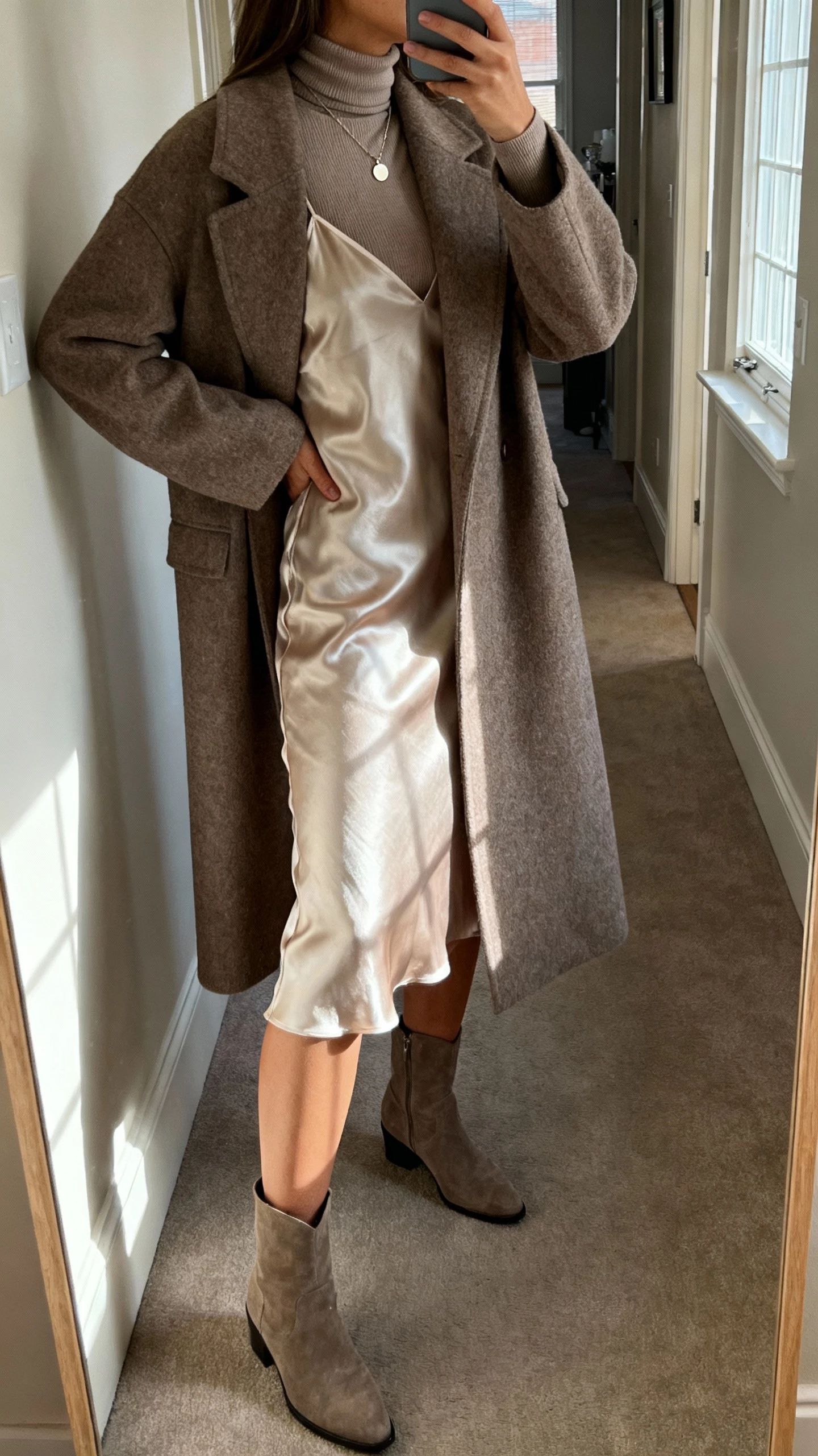 iPhone side-angle selfie of a woman wearing a satin slip dress layered over a fitted turtleneck with a longline wool coat, ankle boots, and simple pendant necklace, face not visible, hallway mirror in an apartment, morning window light, natural iPhone aesthetic.
