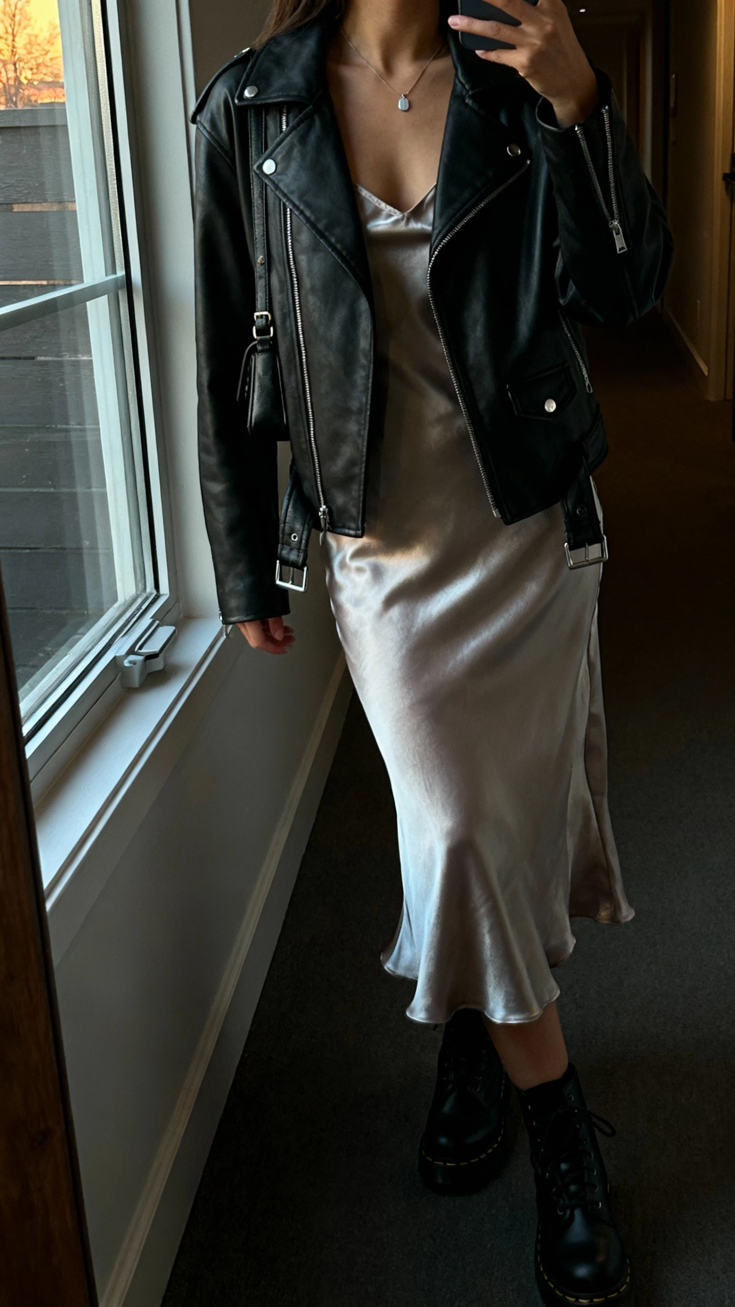 iPhone side-angle selfie of a woman wearing a satin slip dress under a black leather biker jacket, black Doc Martens, minimal pendant necklace, small crossbody bag, face not visible, taken in a dim hallway near a window, evening natural light, casual iPhone aesthetic.