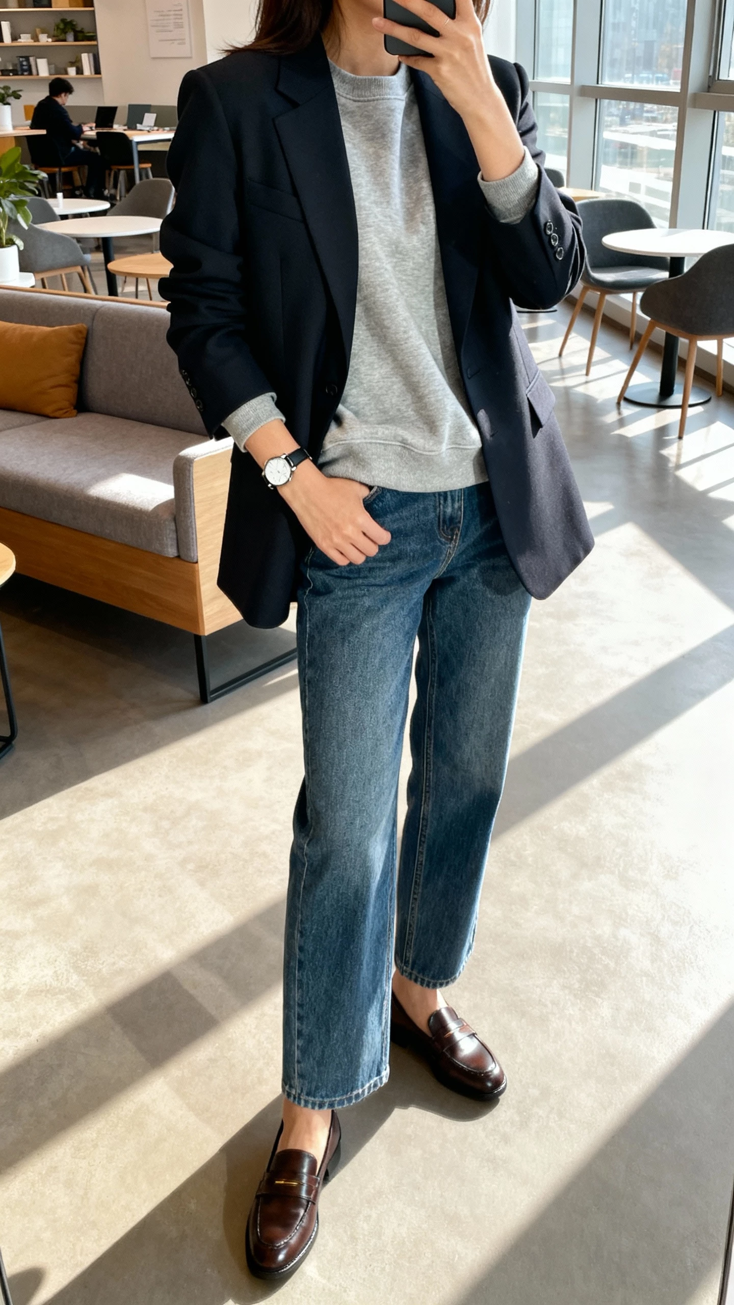 iPhone side-angle selfie of a woman wearing a tailored blazer over a crewneck sweatshirt with straight-leg denim, loafers, simple watch, face not visible, coworking space lounge, morning window light, natural iPhone quality