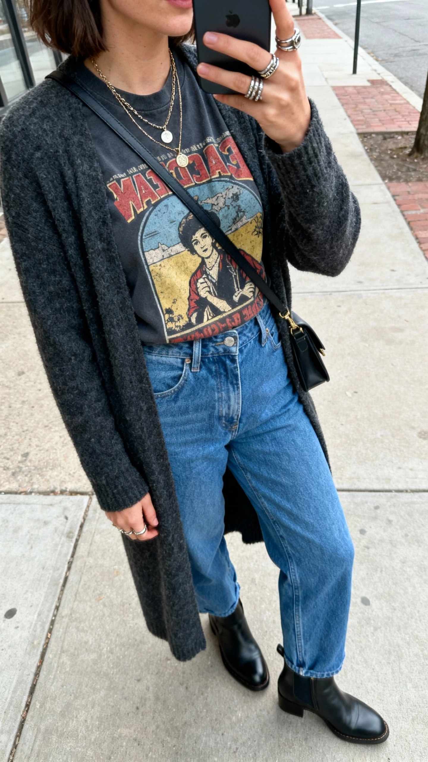 iPhone side-angle selfie of a woman wearing a vintage-inspired graphic tee, blue relaxed straight jeans, longline cardigan, Chelsea ankle boots, layered necklaces, stacked rings, crossbody bag; urban sidewalk, face not visible, natural daylight, natural iPhone photo quality.