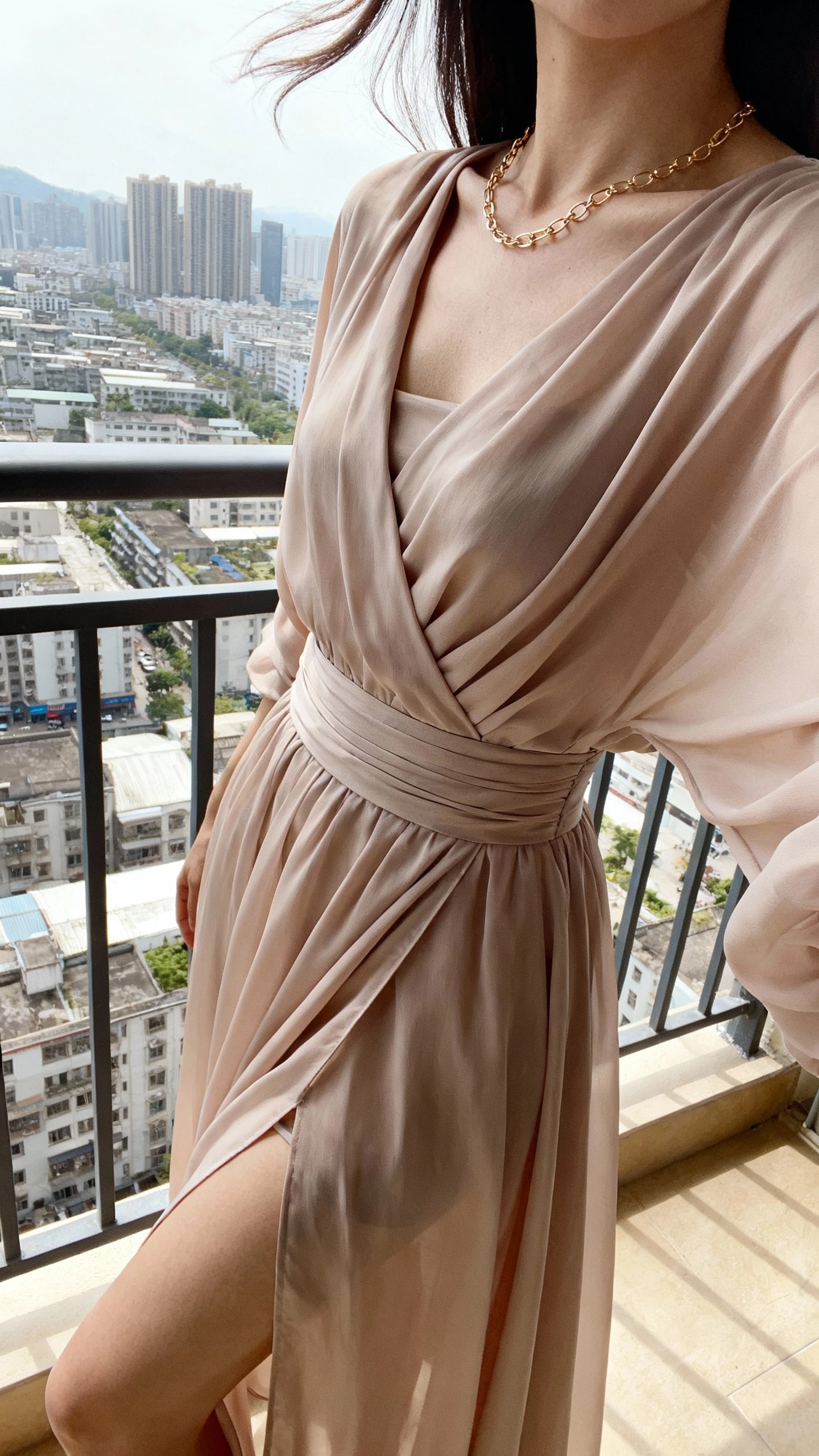 iPhone side-angle selfie of a woman wearing a wrap-style chiffon gown with a high slit and defined waist, delicate chain necklace, face not visible, city balcony setting, breezy daylight, natural iPhone photo quality