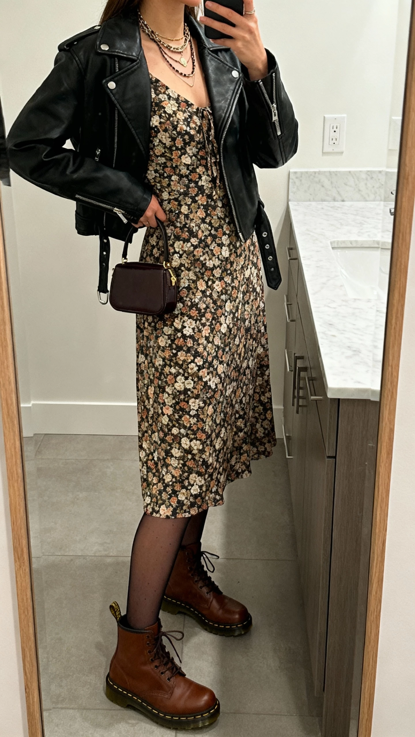 iPhone side-angle selfie of a woman wearing brown Doc Martens with a floral midi dress, black leather moto jacket, sheer tights, layered necklaces, and a tiny top-handle bag, face not visible, bathroom mirror before heading out, indoor lighting, casual iPhone aesthetic.