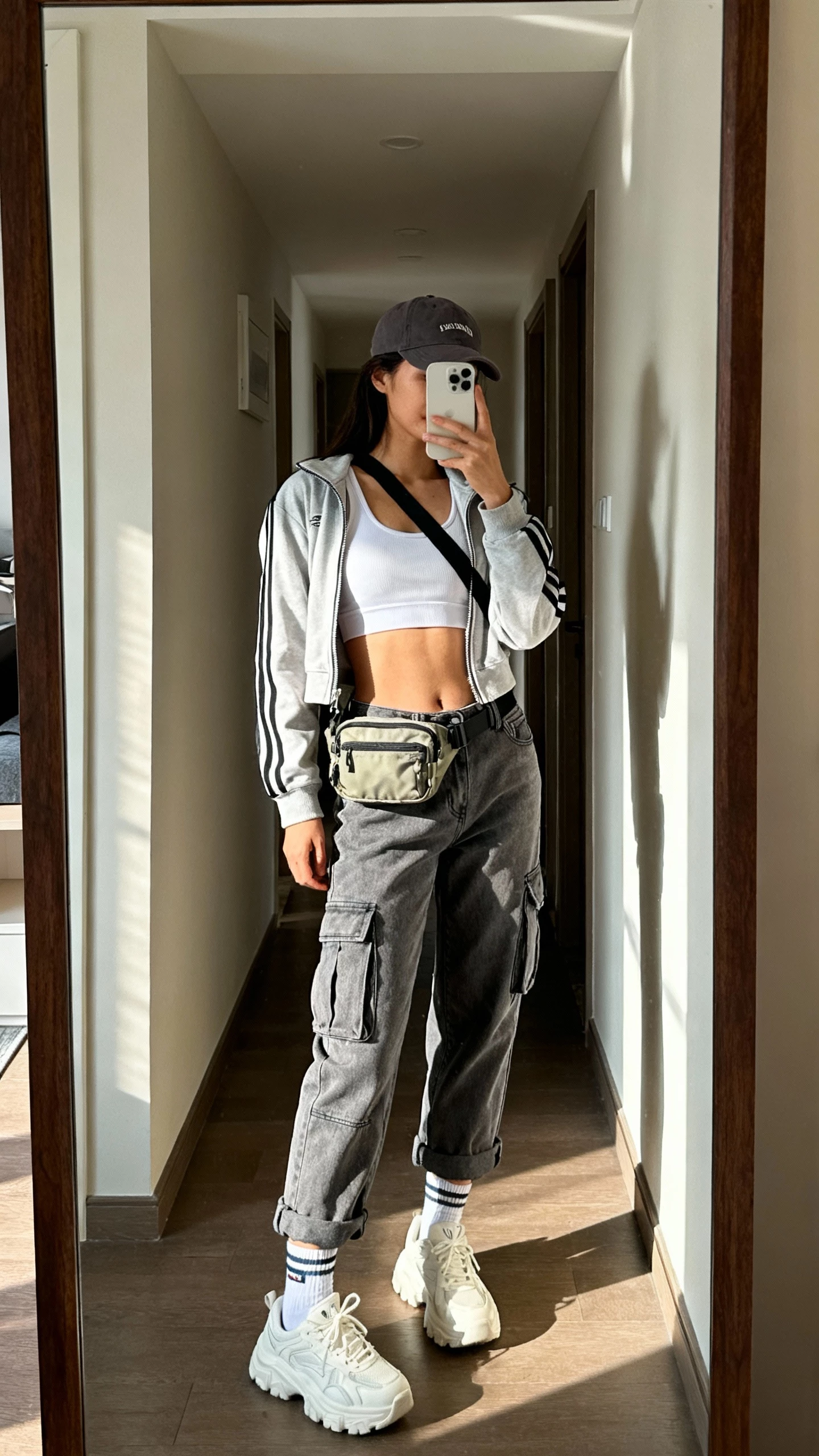 iPhone side-angle selfie of a woman wearing gray cargo jeans, white sport tank, cropped zip-up track jacket, chunky white sneakers, baseball cap, crossbody nylon belt bag, and sporty socks; face not visible, hallway mirror in an apartment, morning daylight, casual iPhone photo.