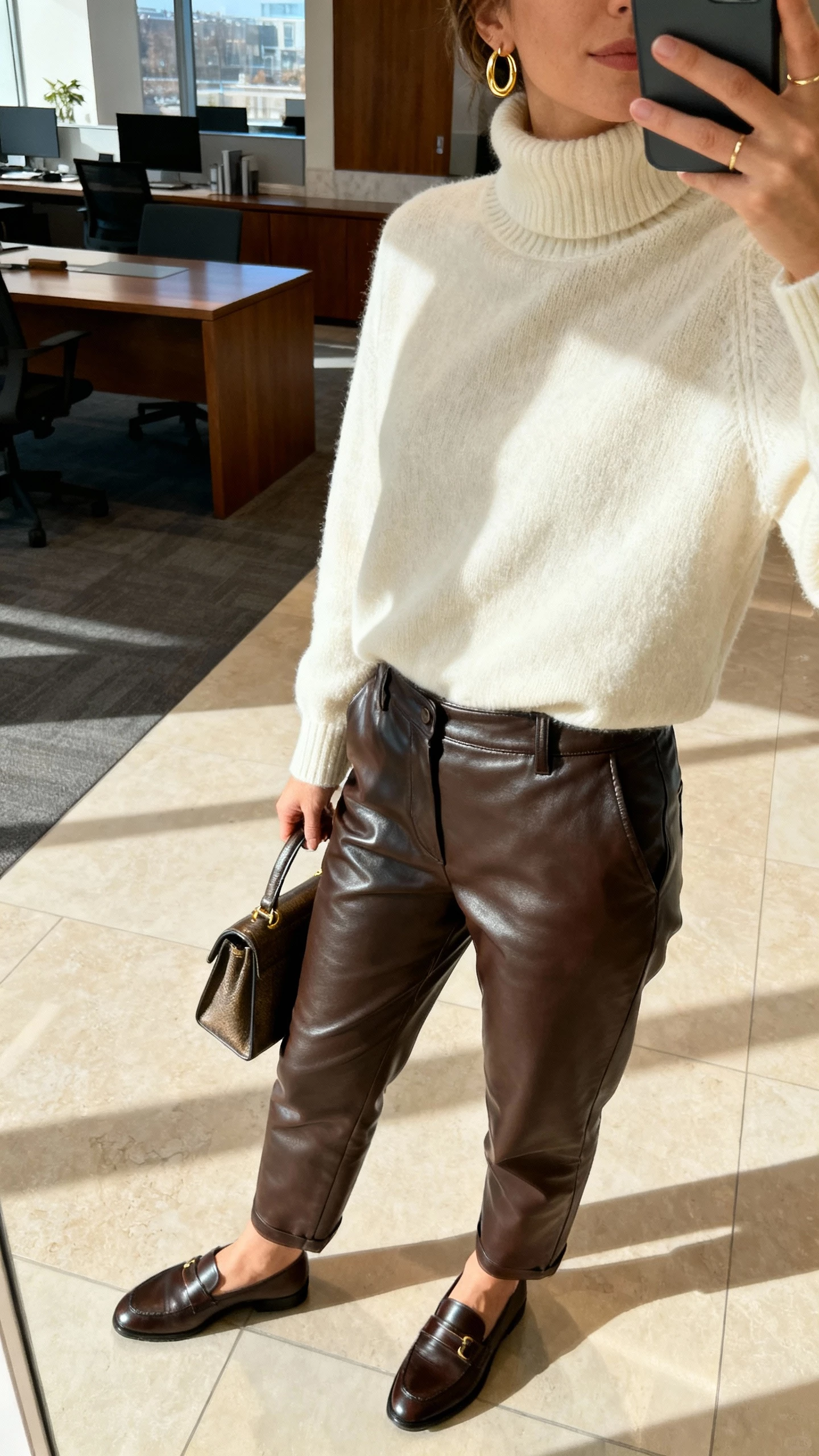 iPhone side-angle selfie of a woman wearing high-waisted leather trousers, a cream cozy turtleneck, loafers, delicate gold hoops, and a structured handbag, face not visible, office lobby with morning light, natural iPhone photo quality