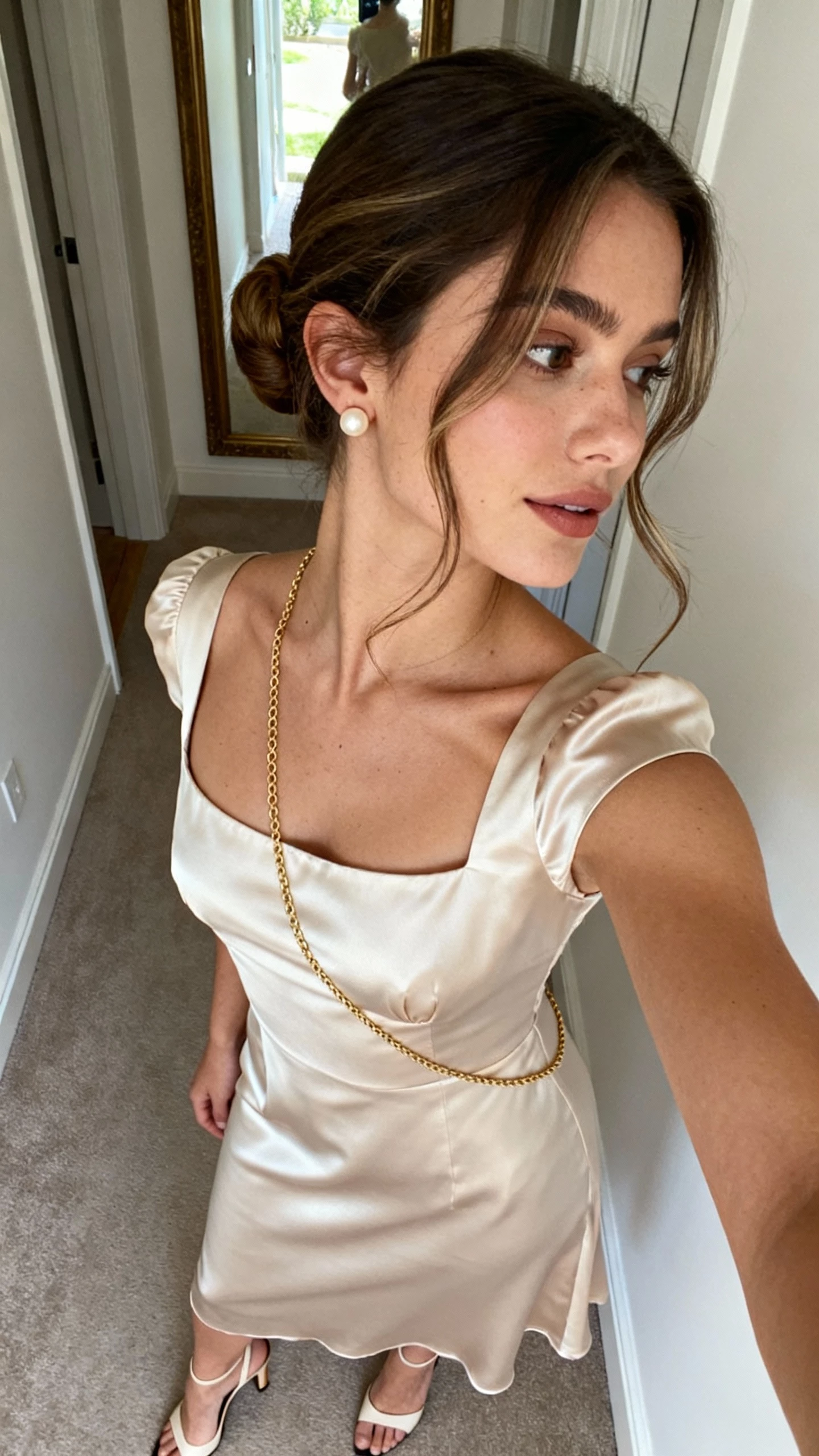 iPhone side-angle selfie of a woman with a French-girl chignon and soft face-framing tendrils, square-neck silk midi dress, delicate pearl studs, thin gold chain, kitten-heel slingbacks; face not visible, hallway mirror, afternoon natural light, casual iPhone aesthetic.