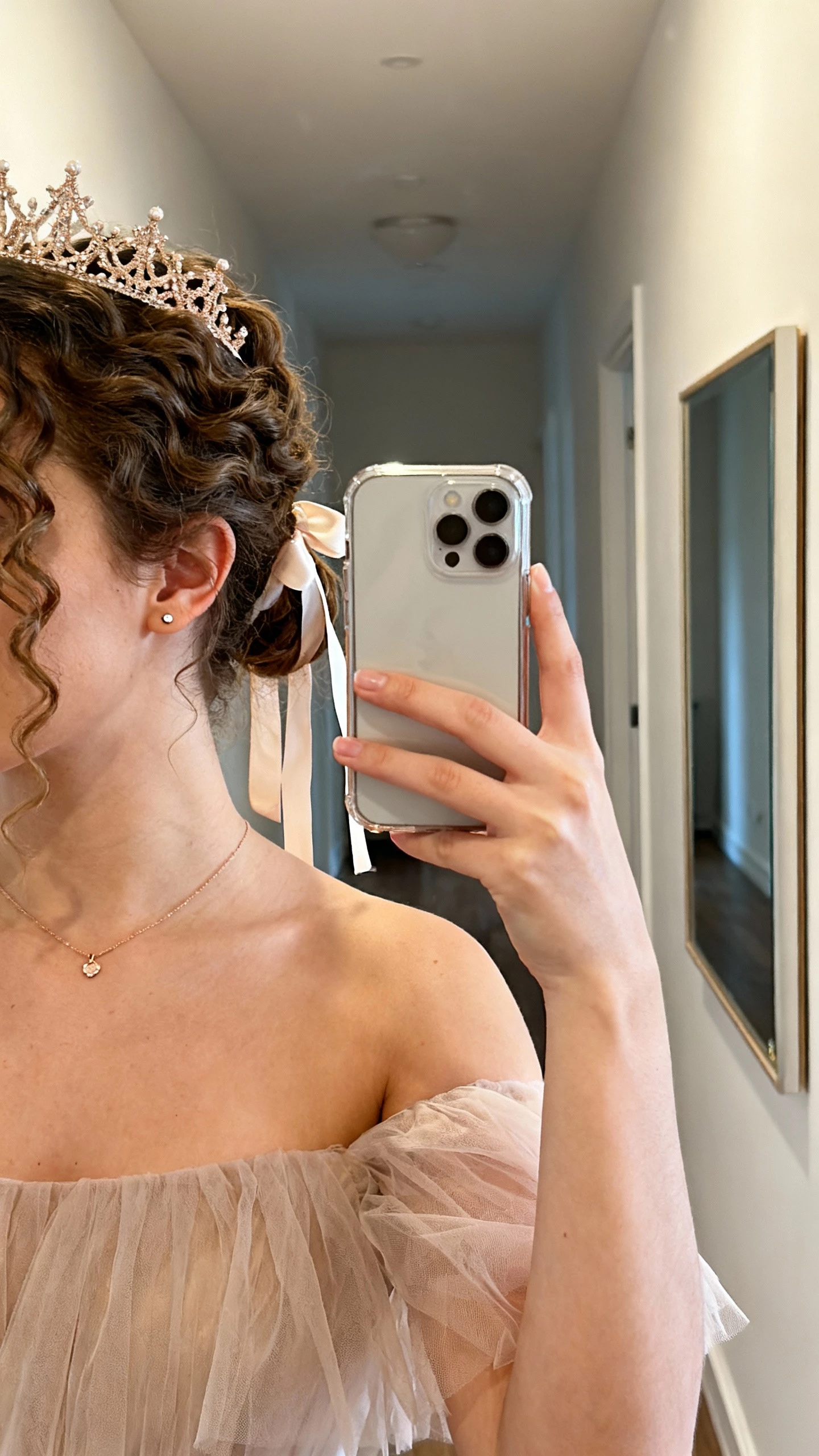 iPhone side-angle selfie of a woman with a half-up curly crown tied with a soft ribbon, off-the-shoulder tulle dress, dainty pendant necklace, small stud earrings, face not visible, hallway mirror, warm indoor light, natural iPhone photo.