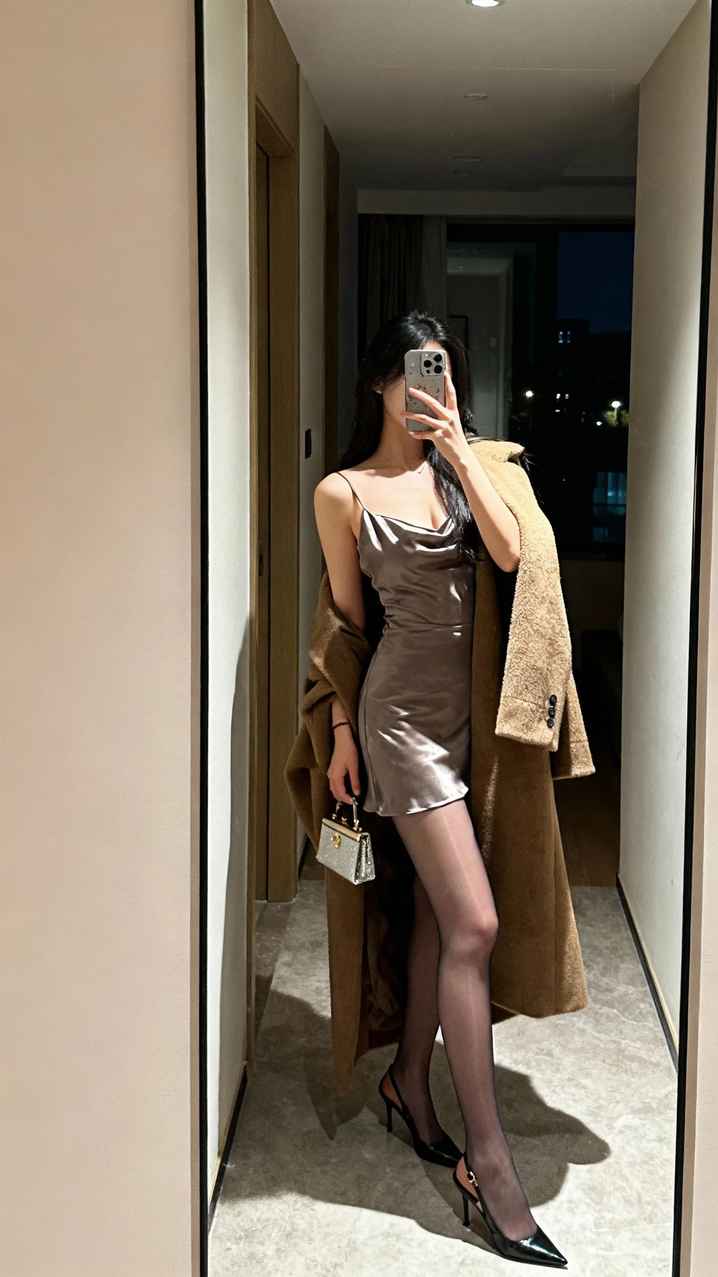 iPhone side-angle selfie showing a woman in a satin slip dress, sheer tights, and a statement coat draped over shoulders, with slingback heels and a small evening bag, face not visible, hallway mirror at night with indoor lighting, natural iPhone quality.